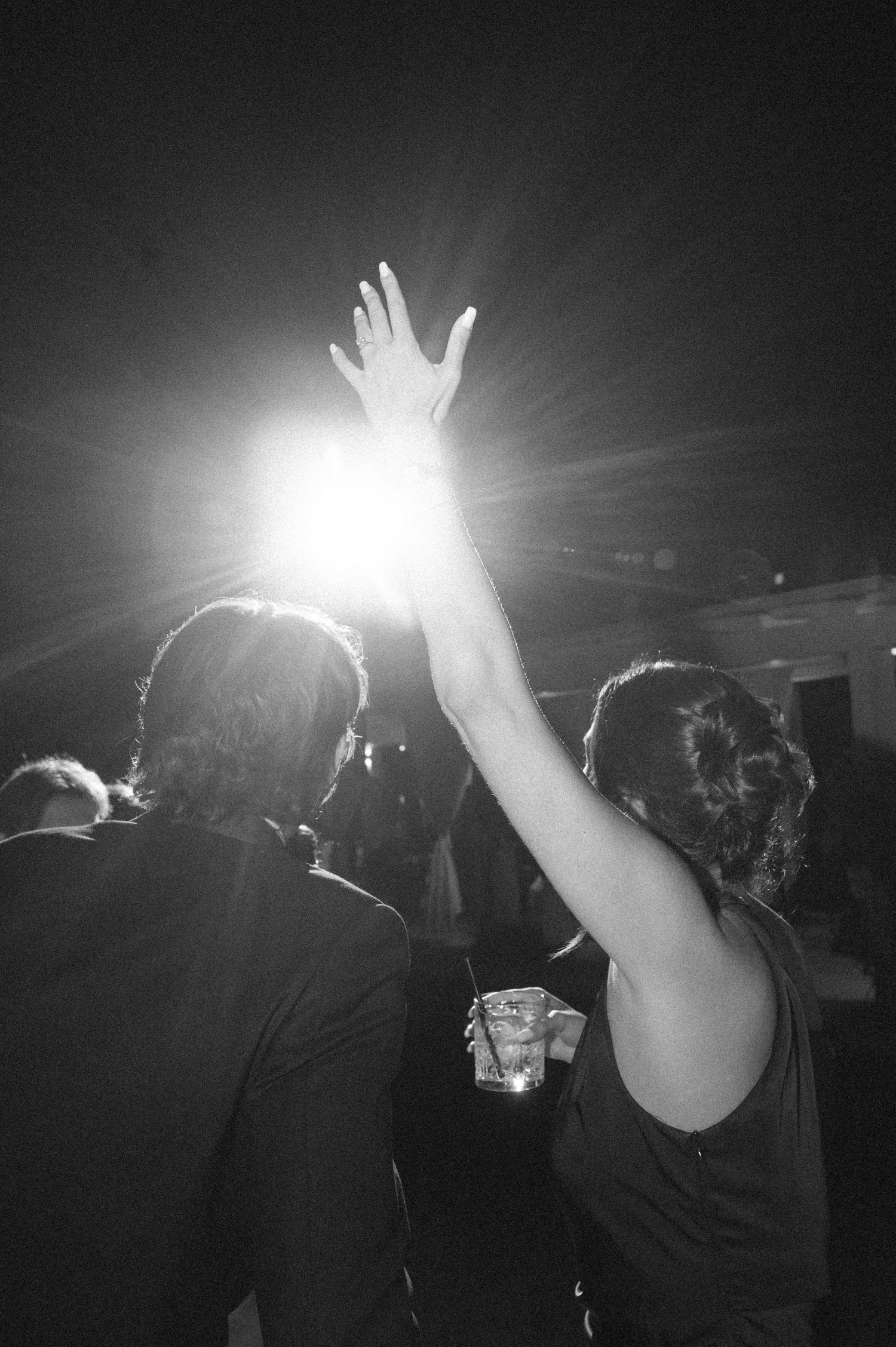 Black and white photo of a woman raising her hand toward a bright stage light at a concert or party.