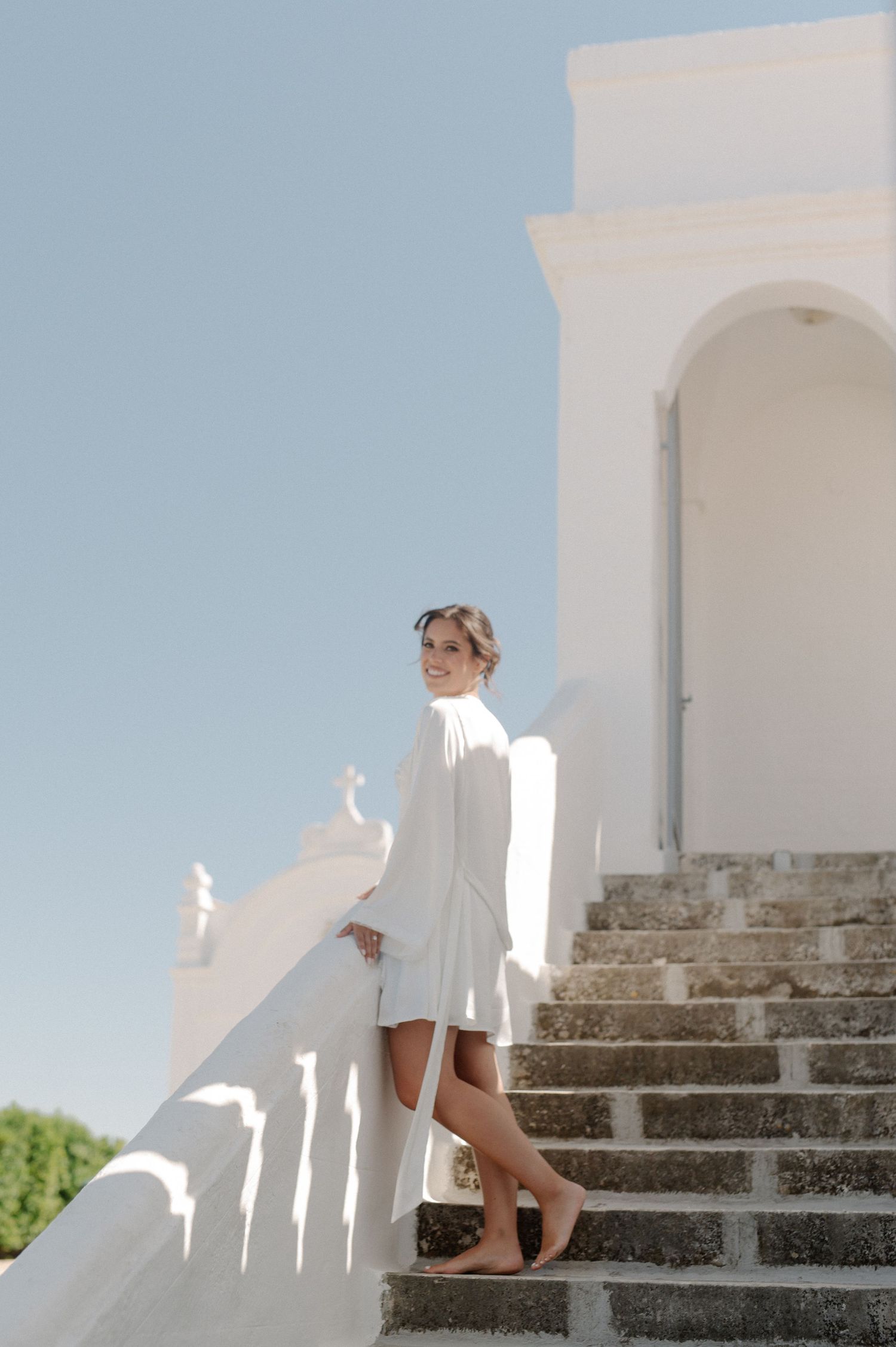 Elegant bride in white dress posing on stone steps of a white church on a sunny day.