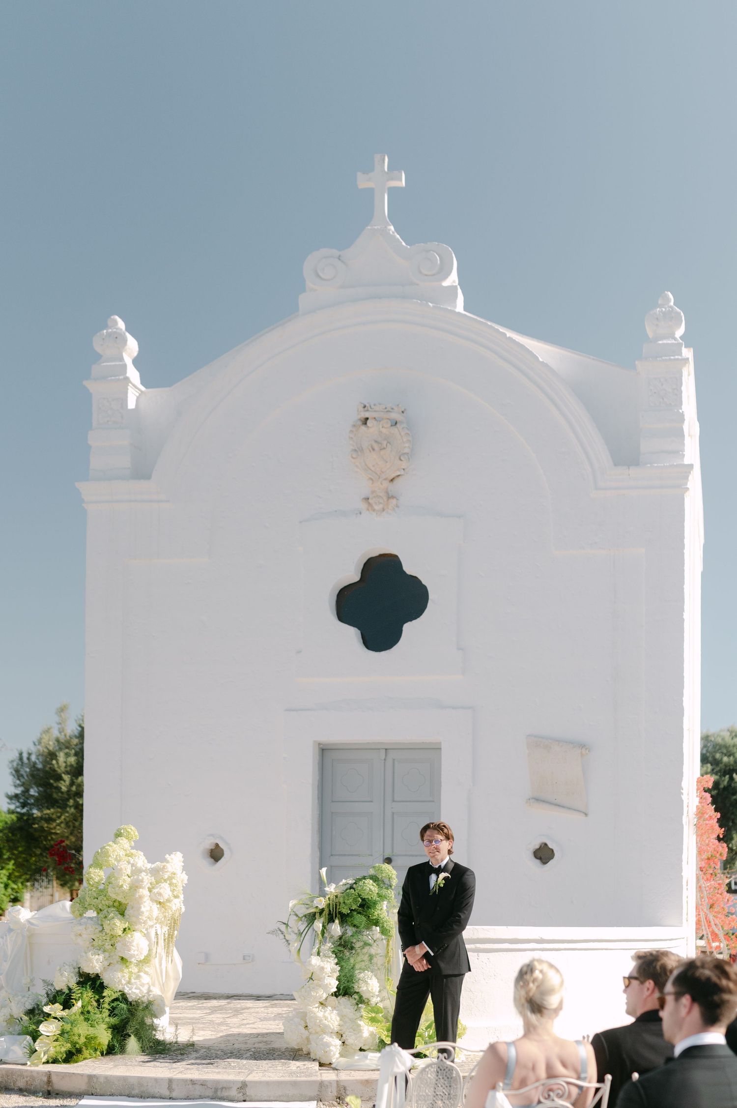 Groom stands before a white Mediterranean chapel with floral arrangements at an outdoor wedding ceremony.