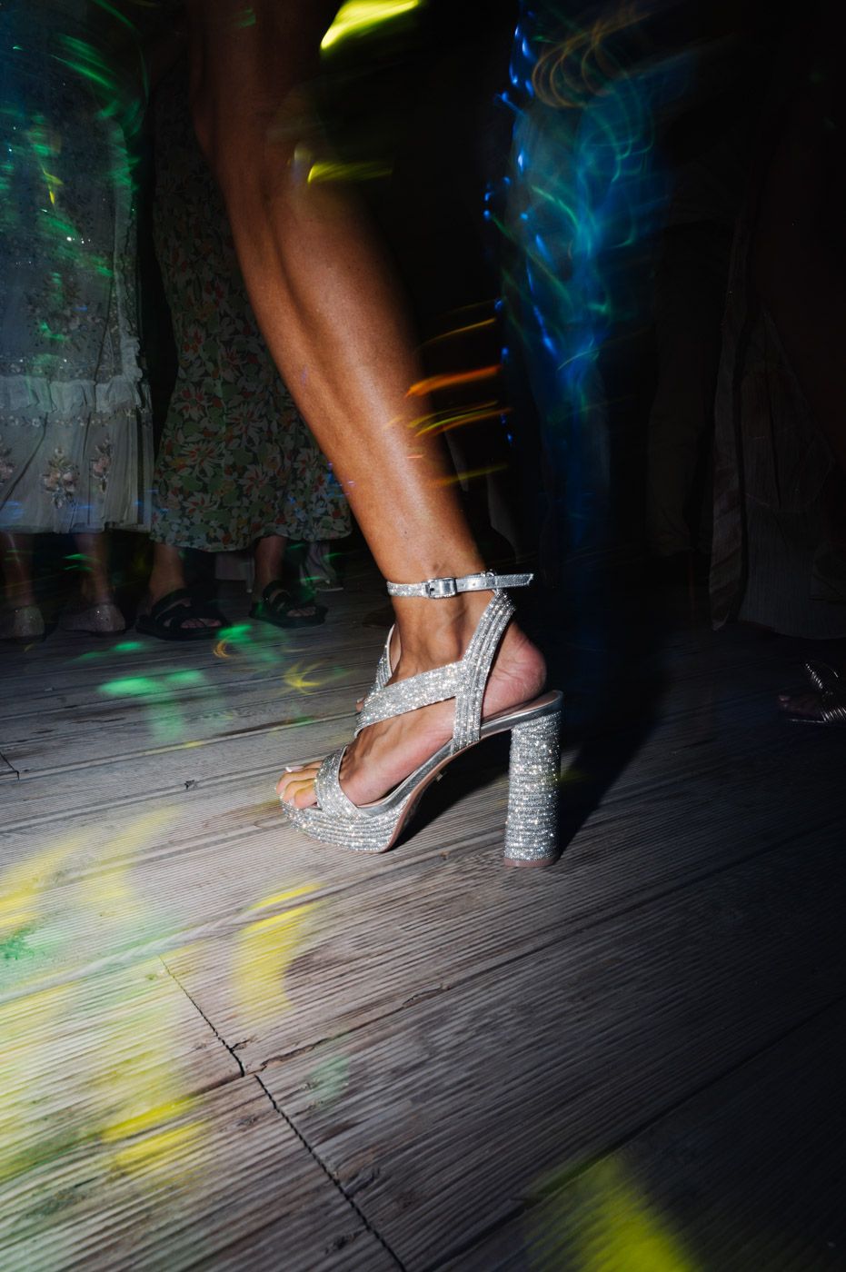 Close-up of sparkly silver heels dancing on wooden floor with colorful lighting.