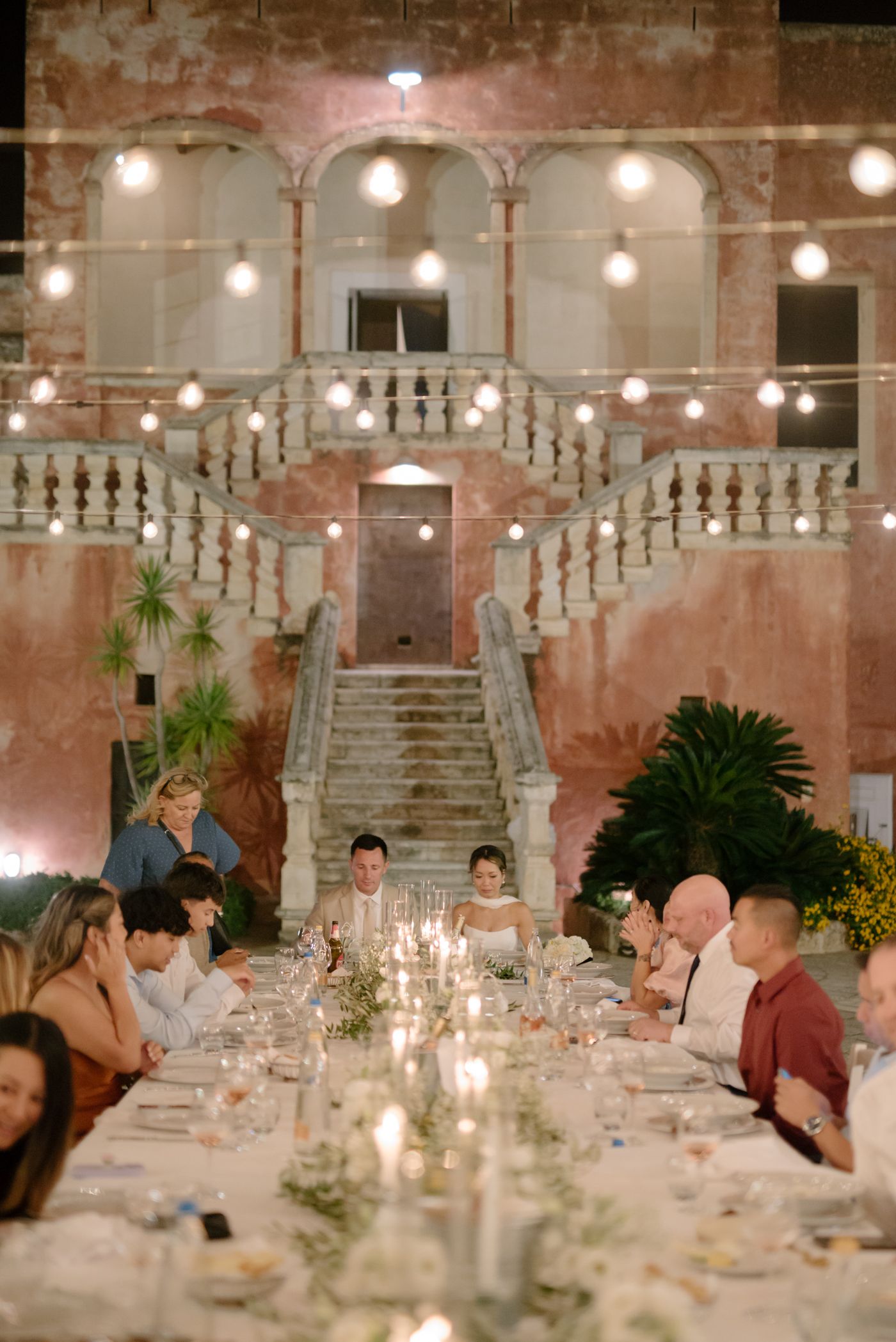 Elegant wedding reception dinner in historic venue with pink walls, marble staircase and string lights creating romantic ambiance.