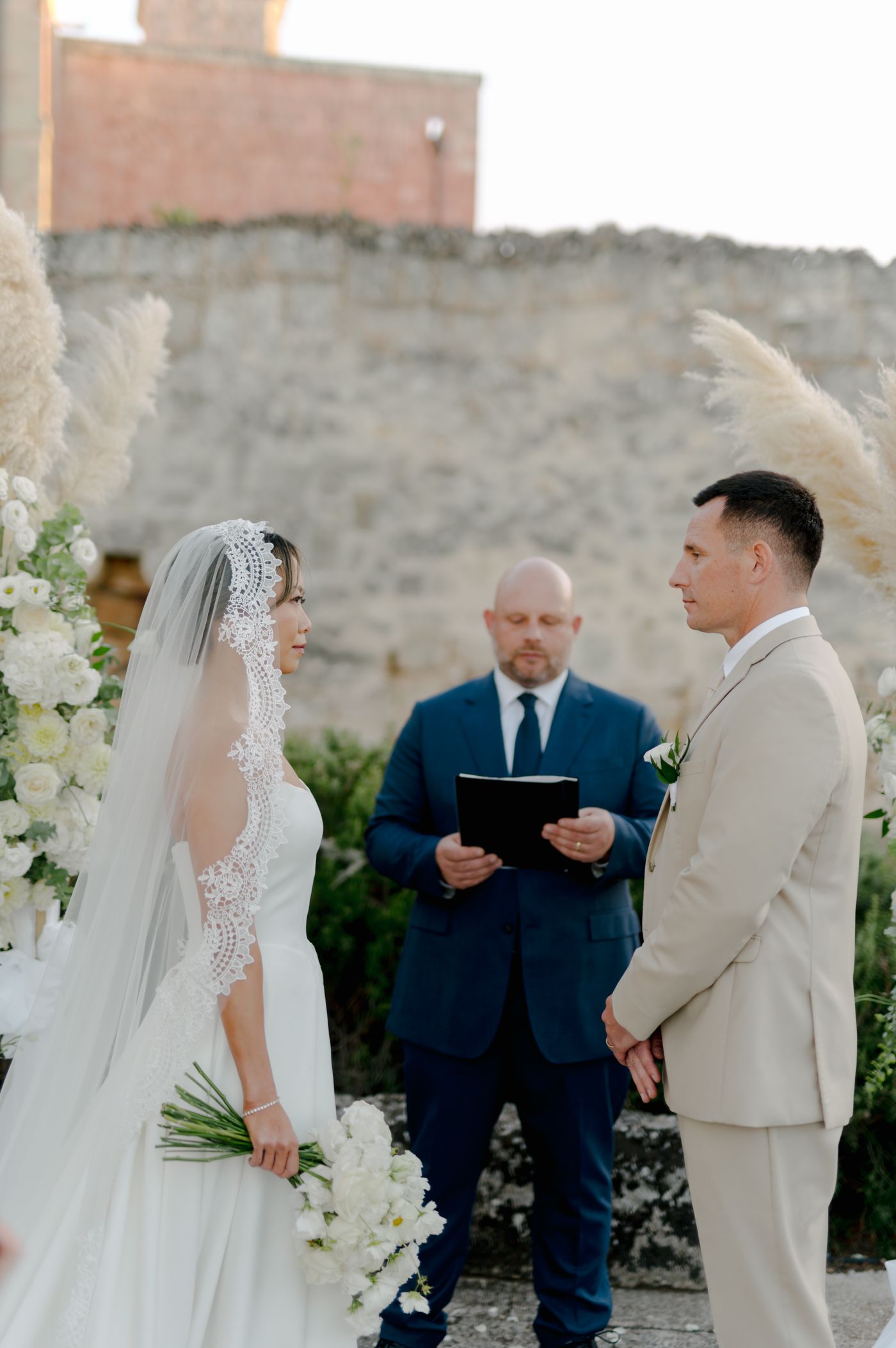 Wedding ceremony taking place outdoors with white floral decorations and an officiant in navy suit.