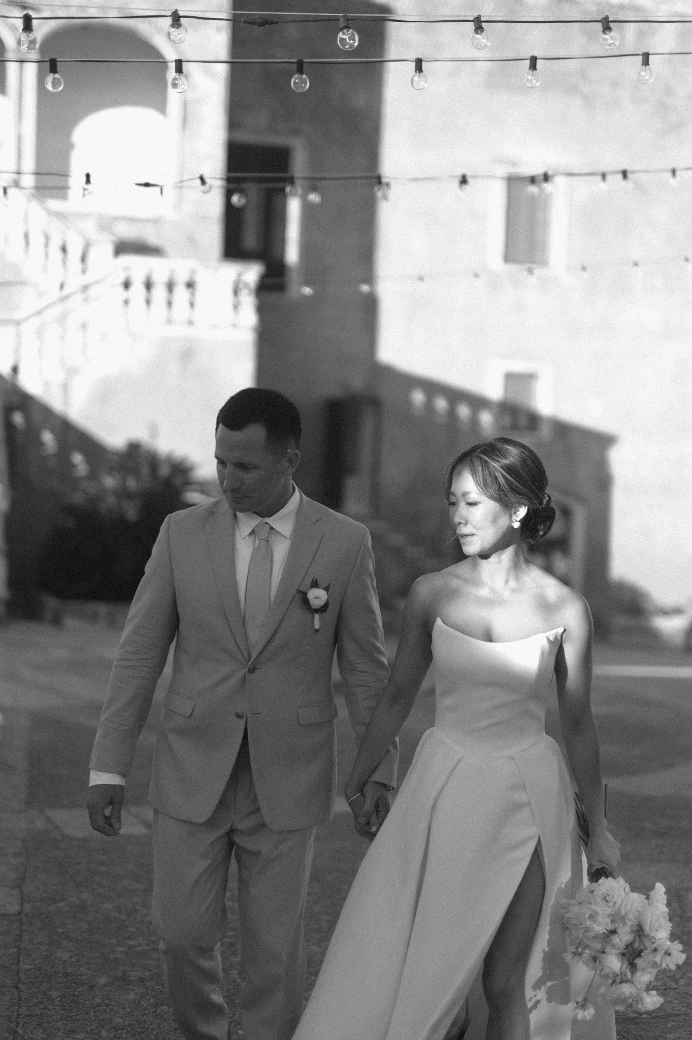Black and white wedding photo of a couple walking together outdoors with string lights and architecture in the background.