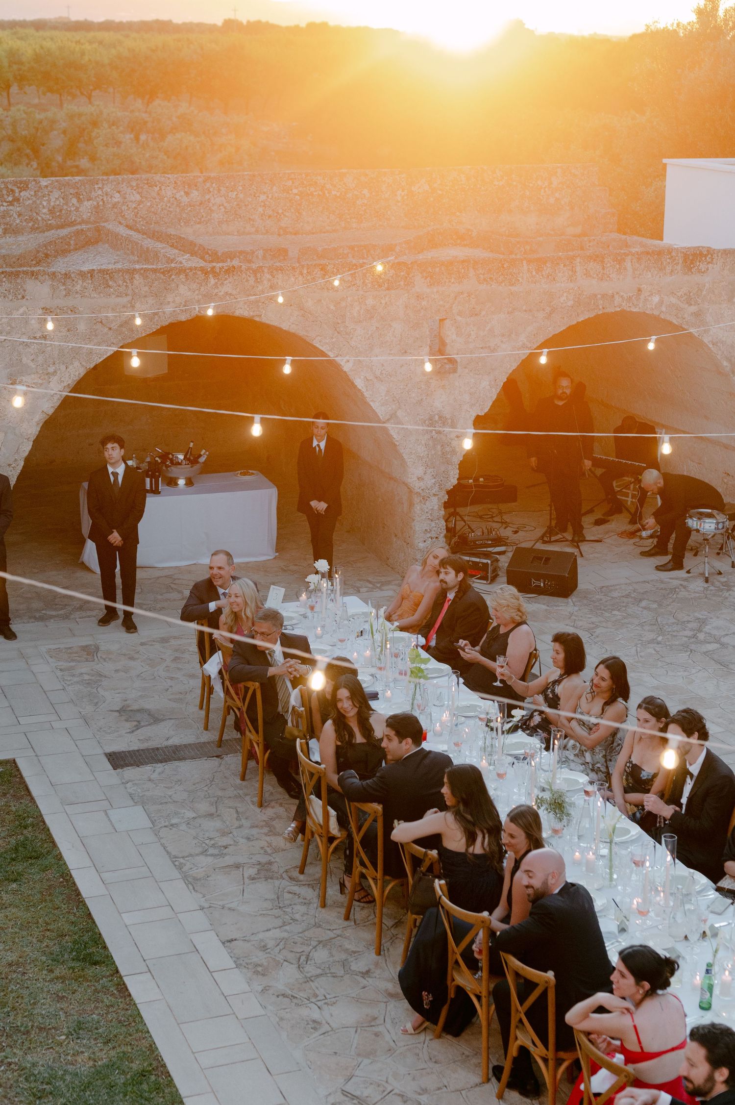 Outdoor wedding reception at sunset with guests seated at long table under string lights beside stone arched wall.