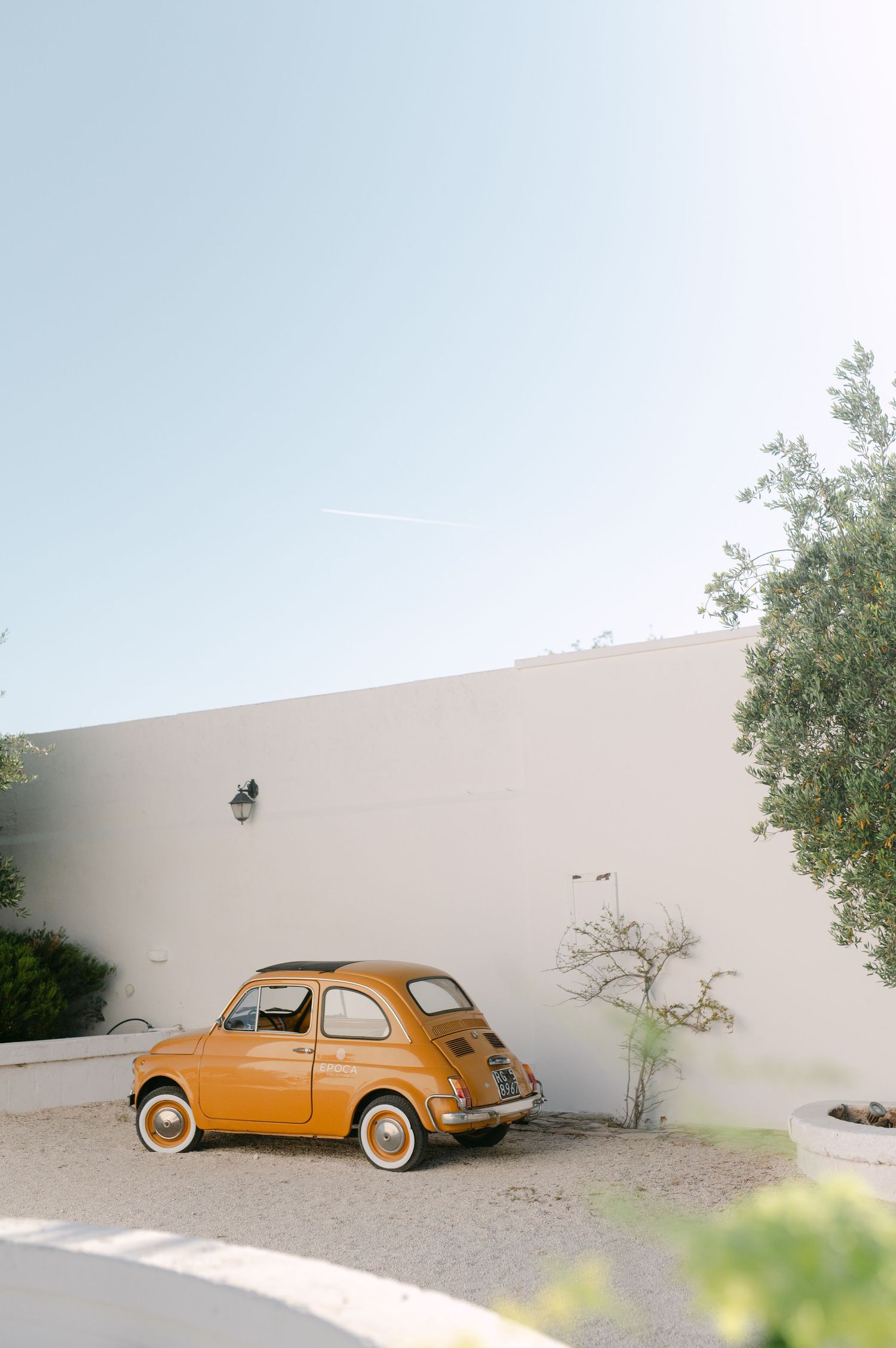 Vintage yellow Fiat 500 parked beside a white stucco wall with a tree in a sunny courtyard.