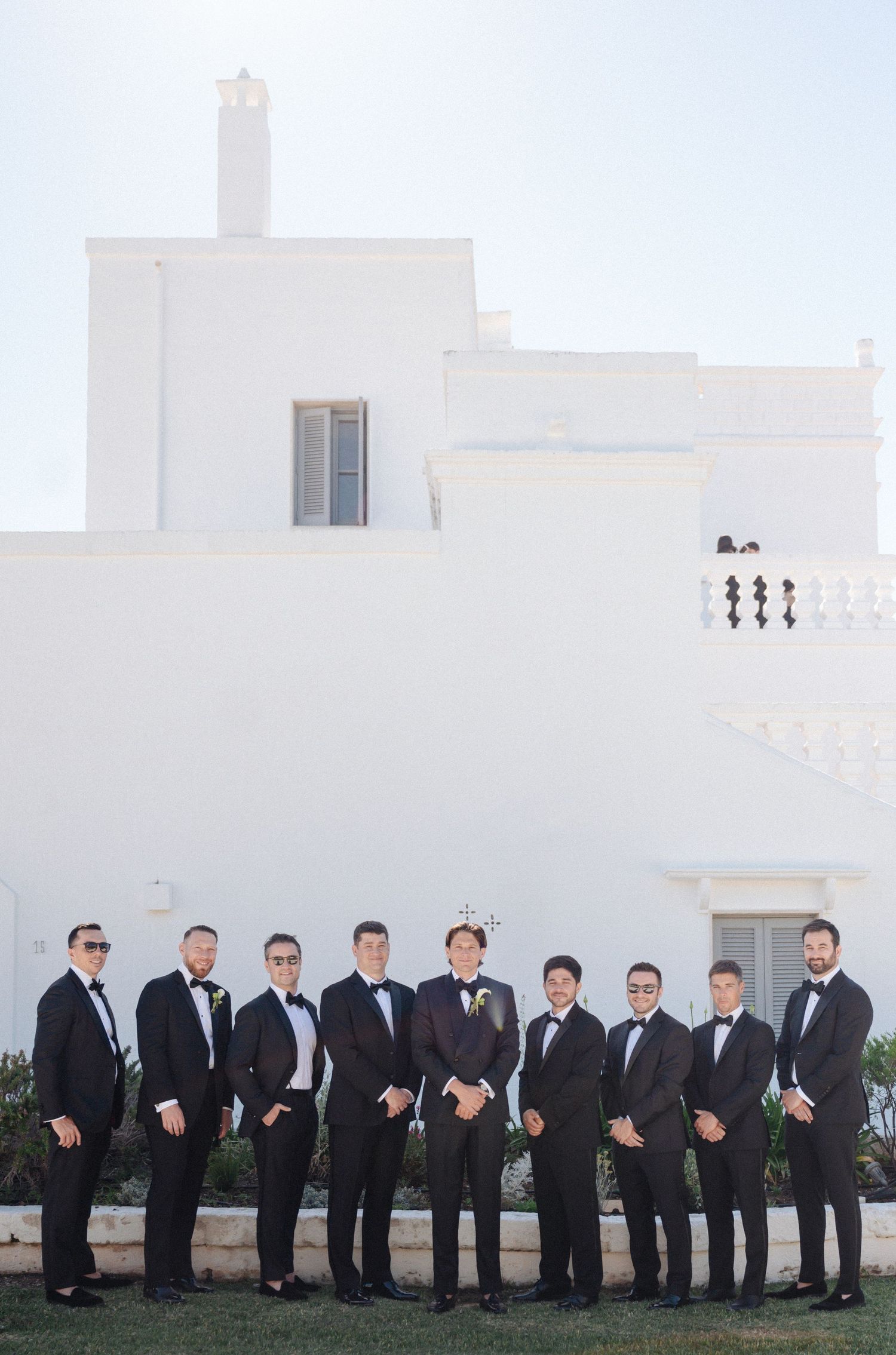 Groom and groomsmen in black tuxedos with bow ties posing in front of a white Mediterranean villa.