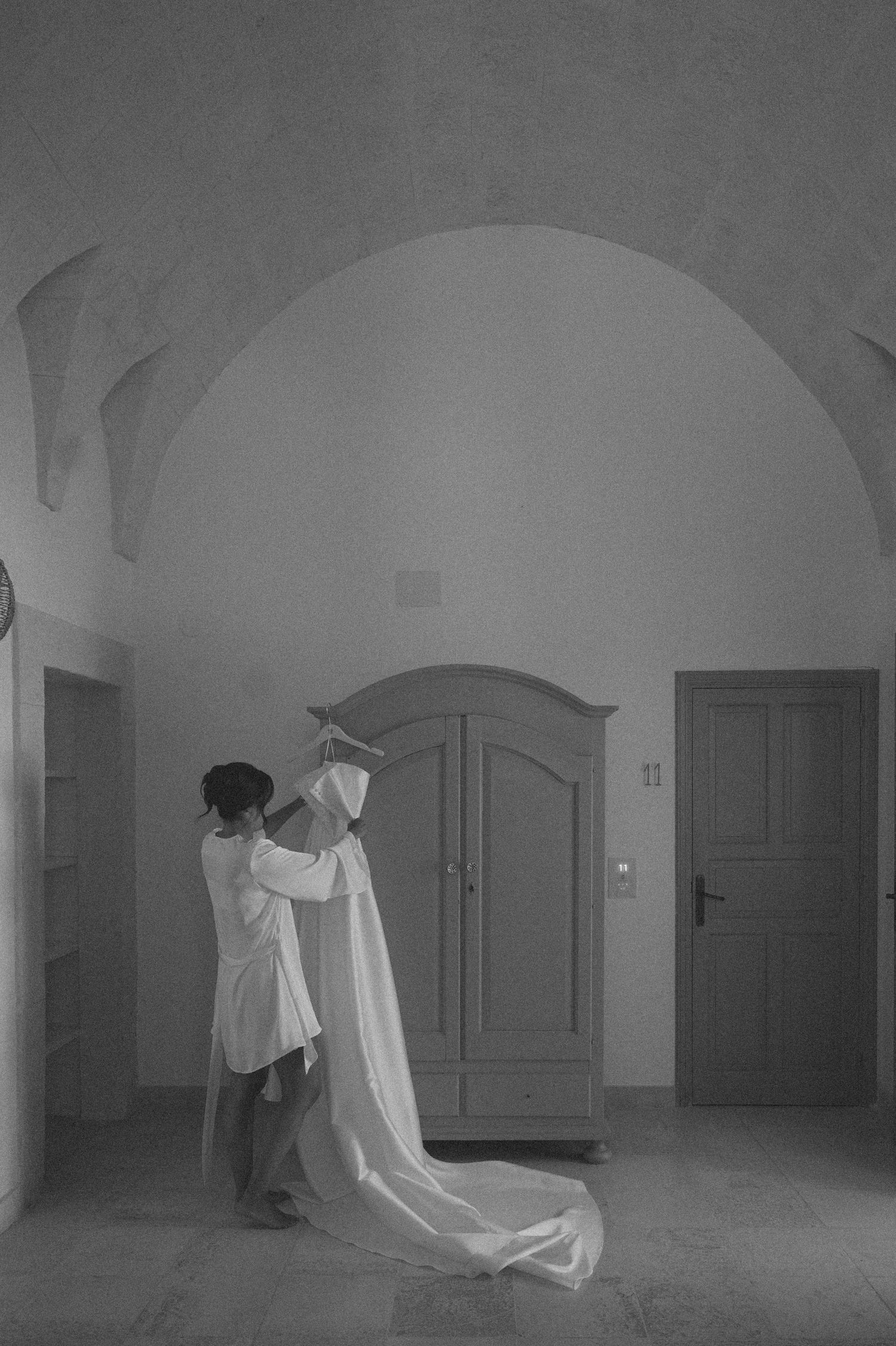 Black and white photo of a bride being helped into her wedding dress in an elegant arched room.
