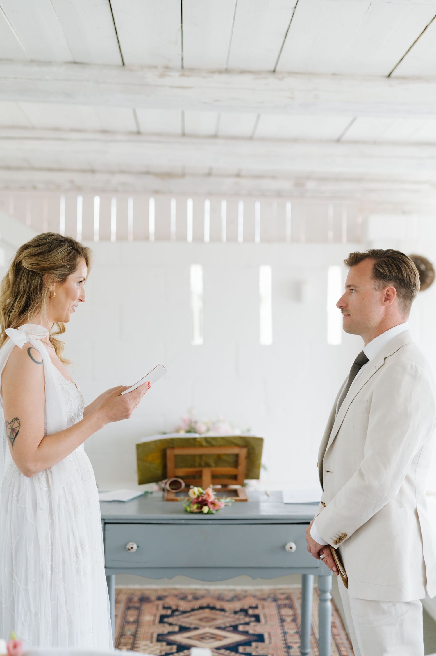 Two people in white attire stand in a bright room with vintage furniture and decor.