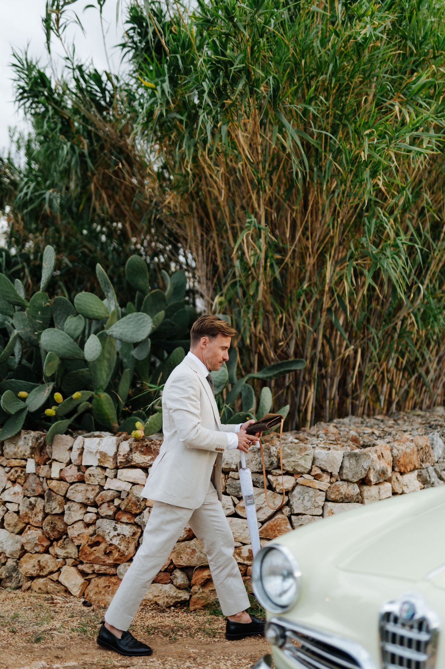 Person in light suit walking alongside vintage cream car with bamboo and cacti background.