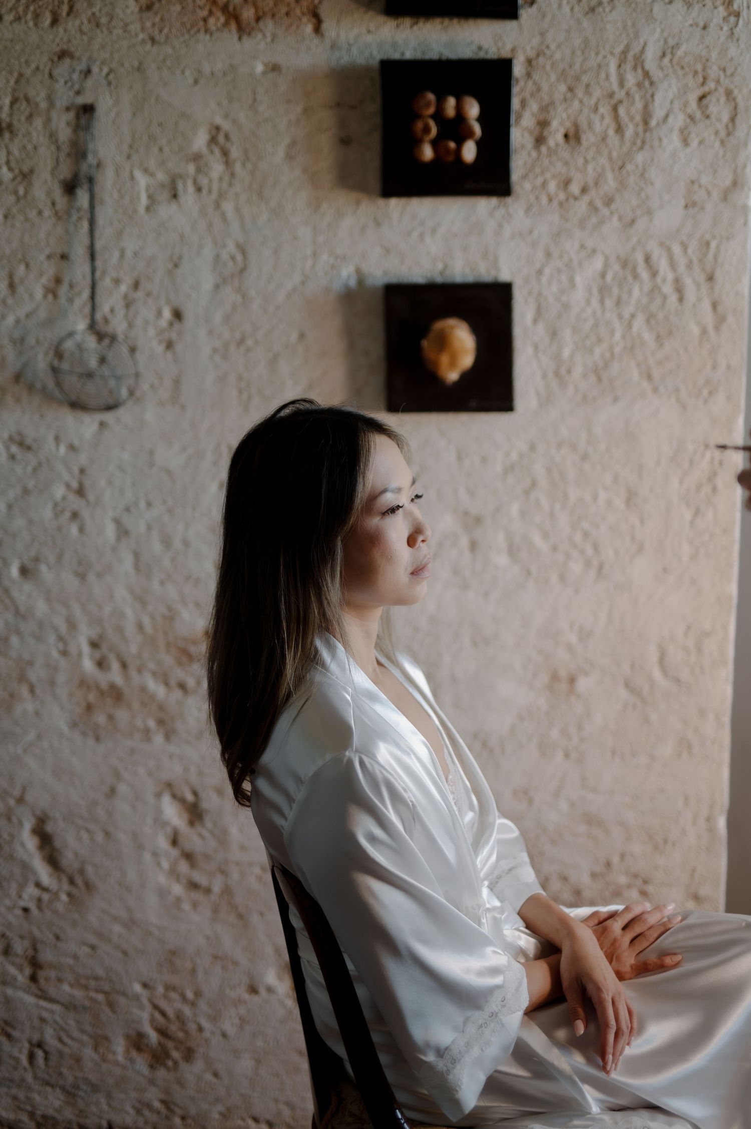 A person in a white silk robe sits contemplatively against a textured wall with framed artwork in the background.