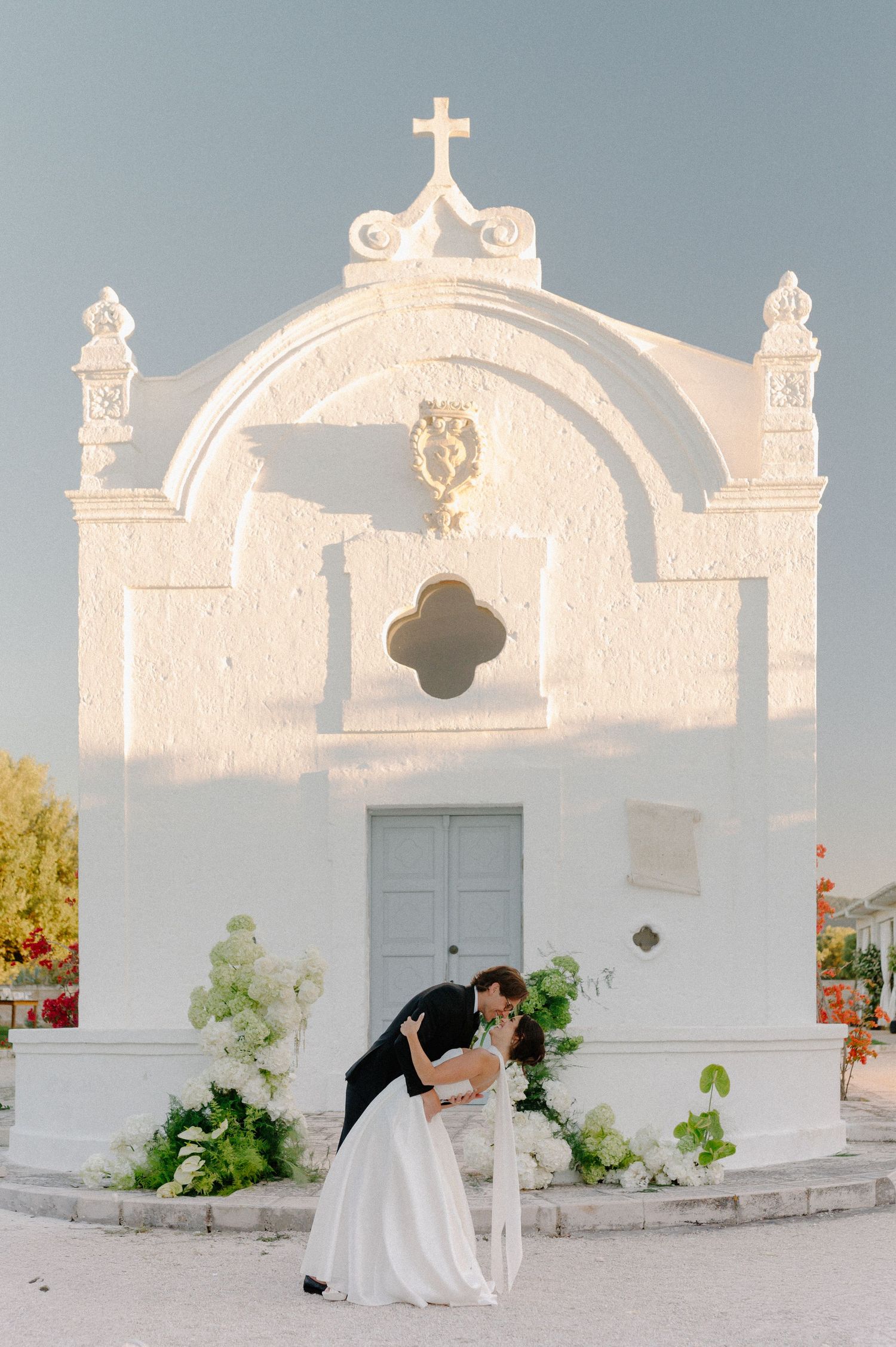 Couple sharing a romantic dip kiss in front of a white Greek chapel adorned with floral arrangements on their wedding day.