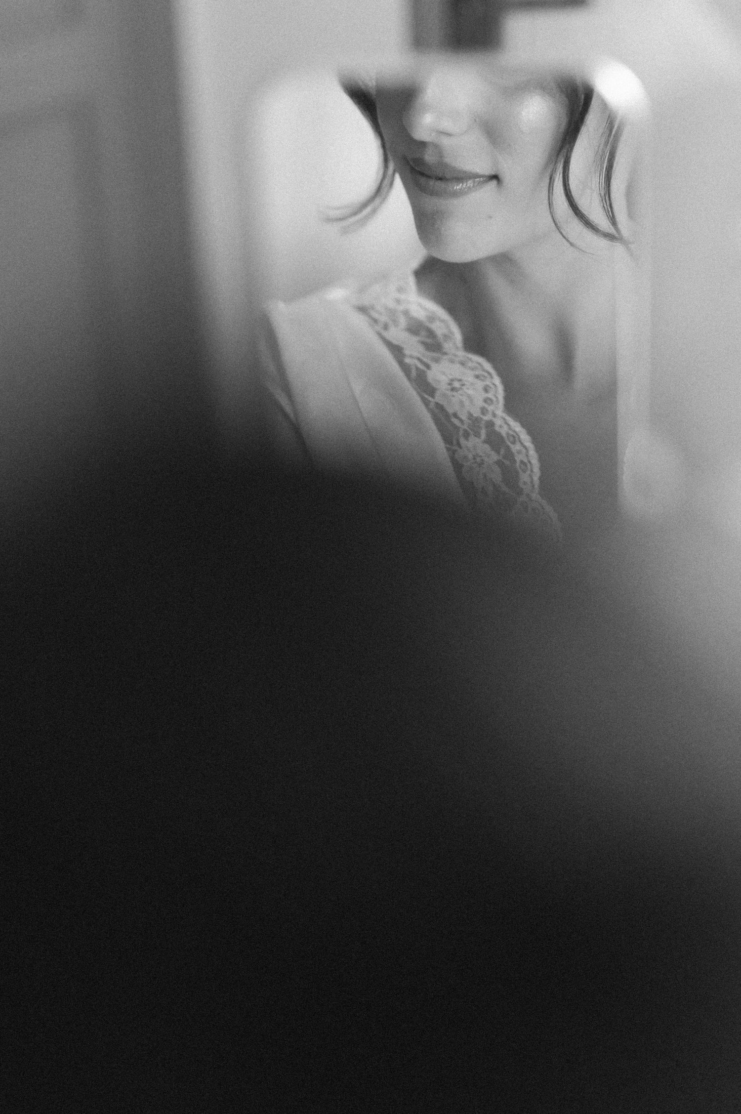 Black and white photo of a smiling bride in lace wedding dress, captured through a soft-focus foreground element.