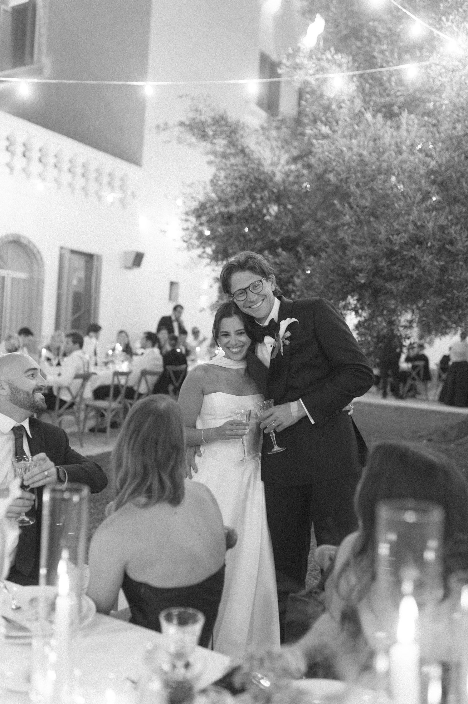 Black and white photo of a smiling bride and groom embracing at their outdoor wedding reception with string lights.