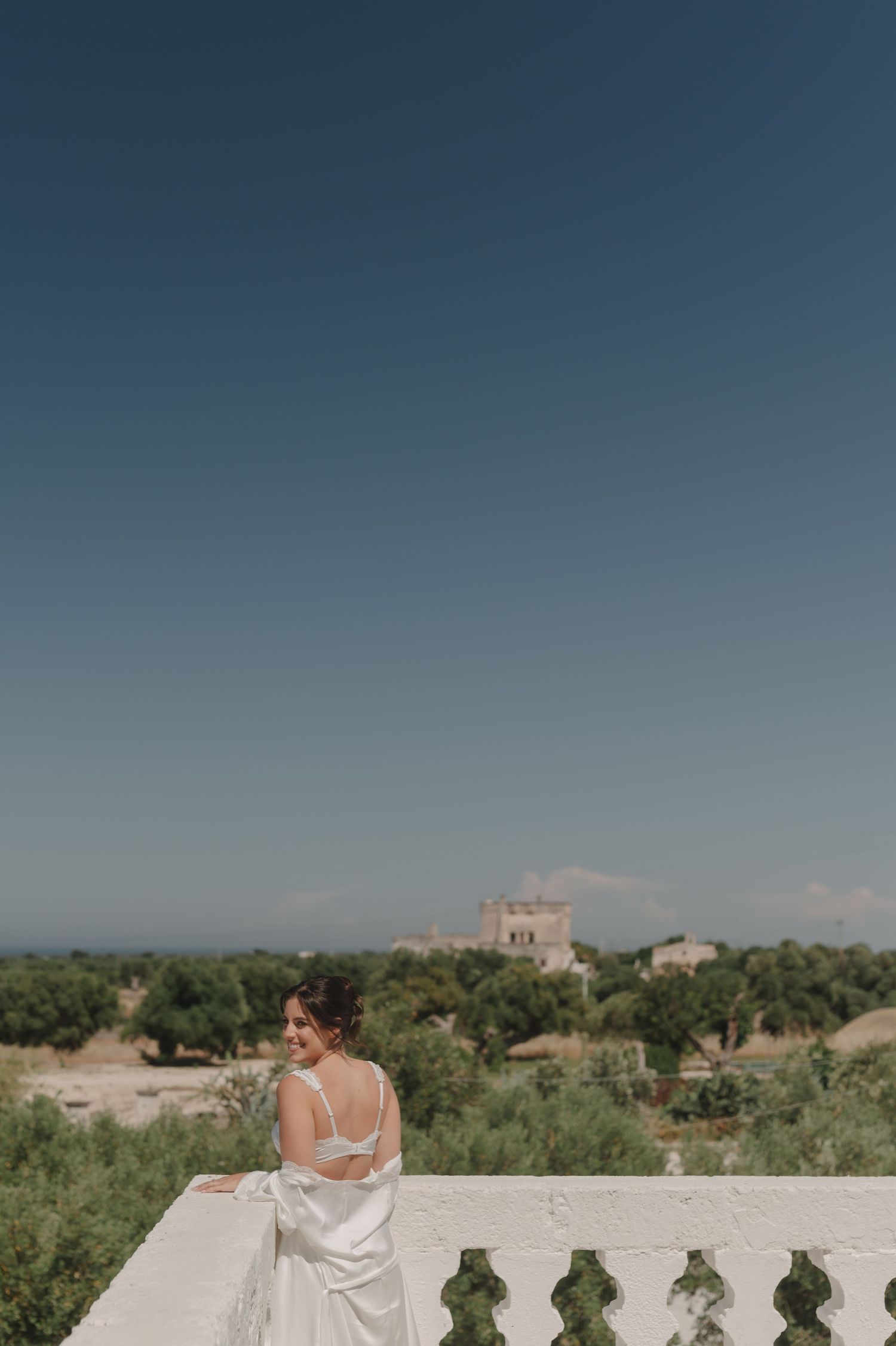 Bride in white gown stands on balcony overlooking lush olive trees and historic villa under clear blue sky.