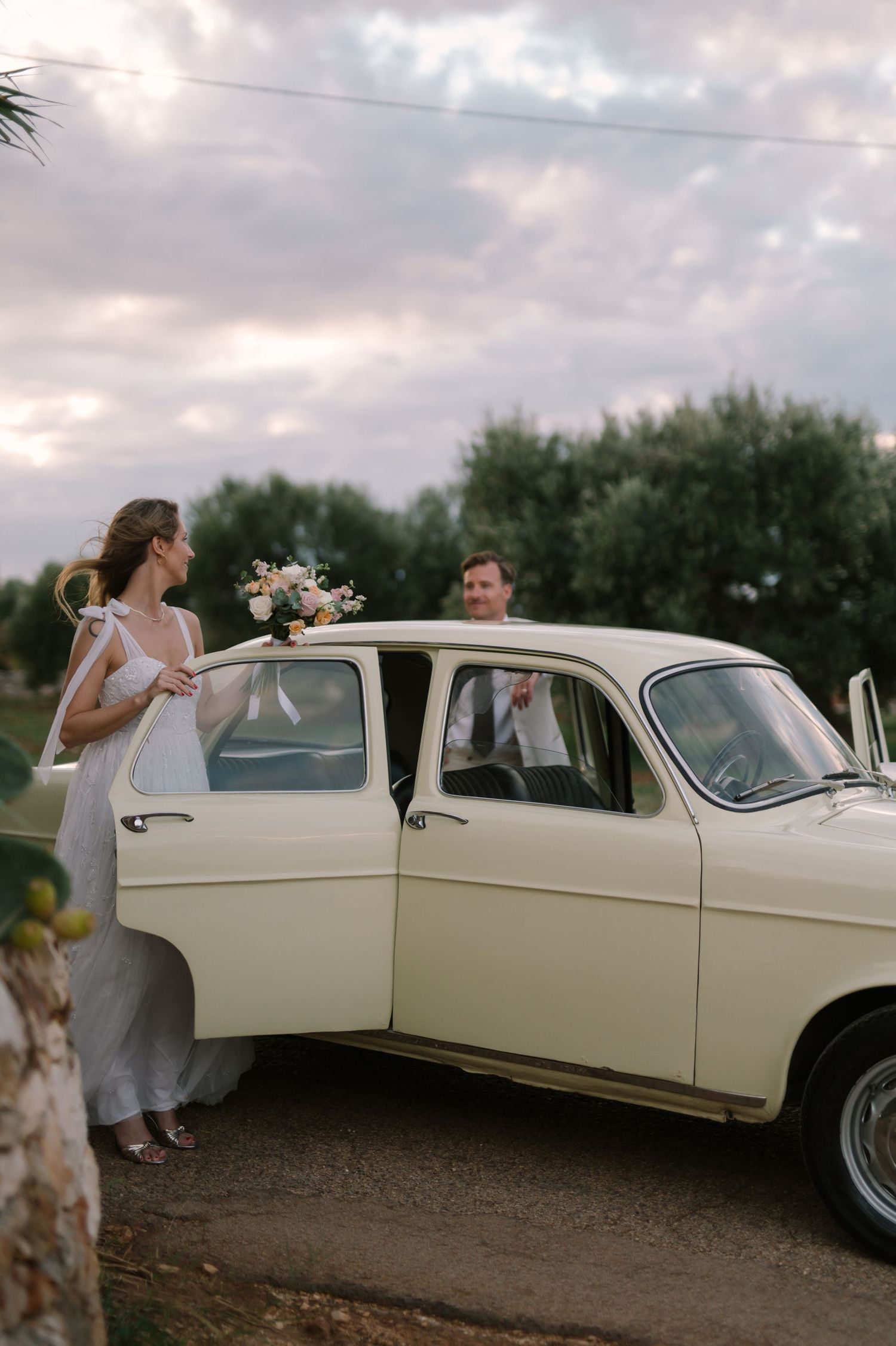 Wedding party gathered around a vintage cream-colored limousine.