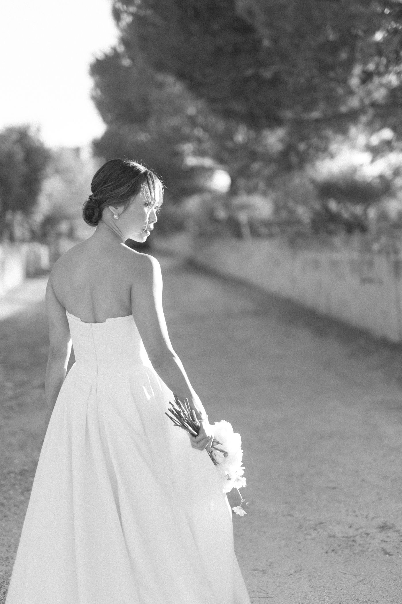 A bride in a strapless white wedding gown holds a bouquet while standing on a path in black and white photography.