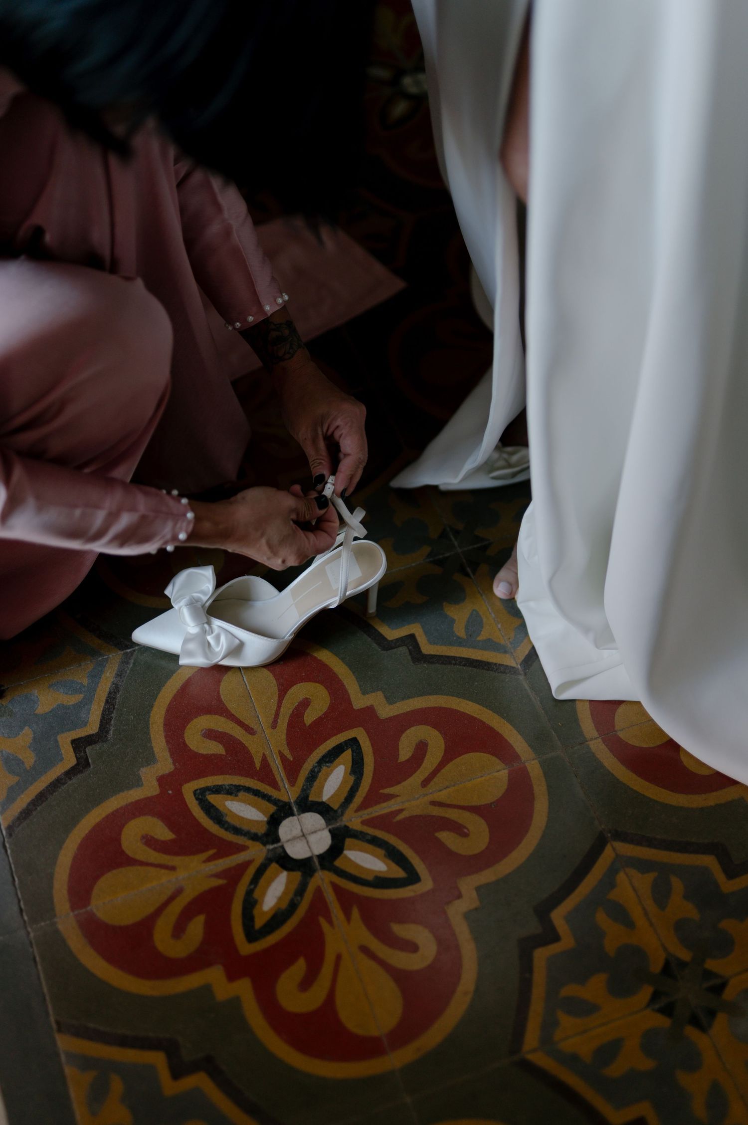 Someone adjusts a white bridal shoe on ornate floral tiled flooring during wedding preparations.