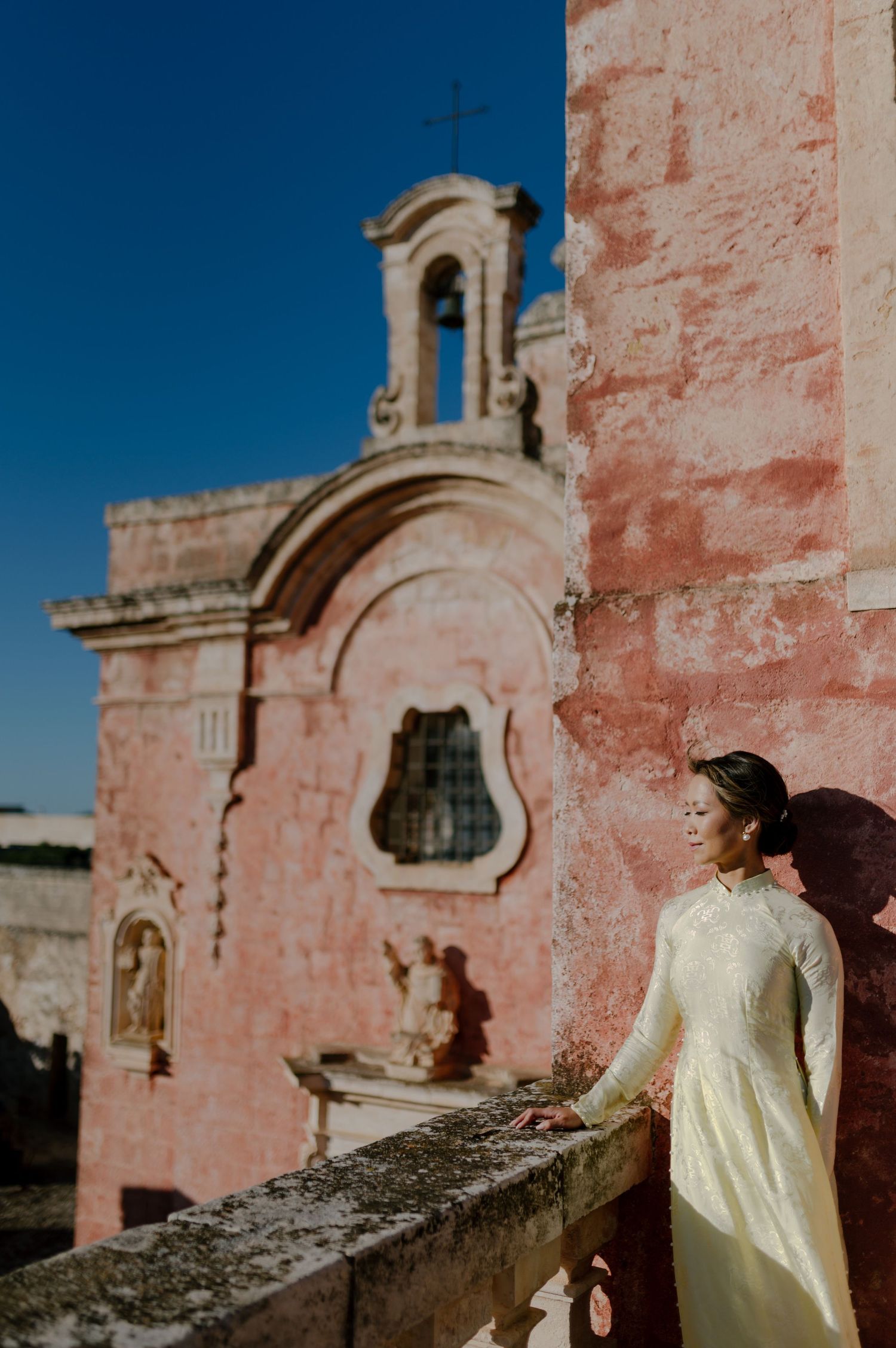 A person in a white dress stands against pink stone walls of an old church or mission building under blue sky.