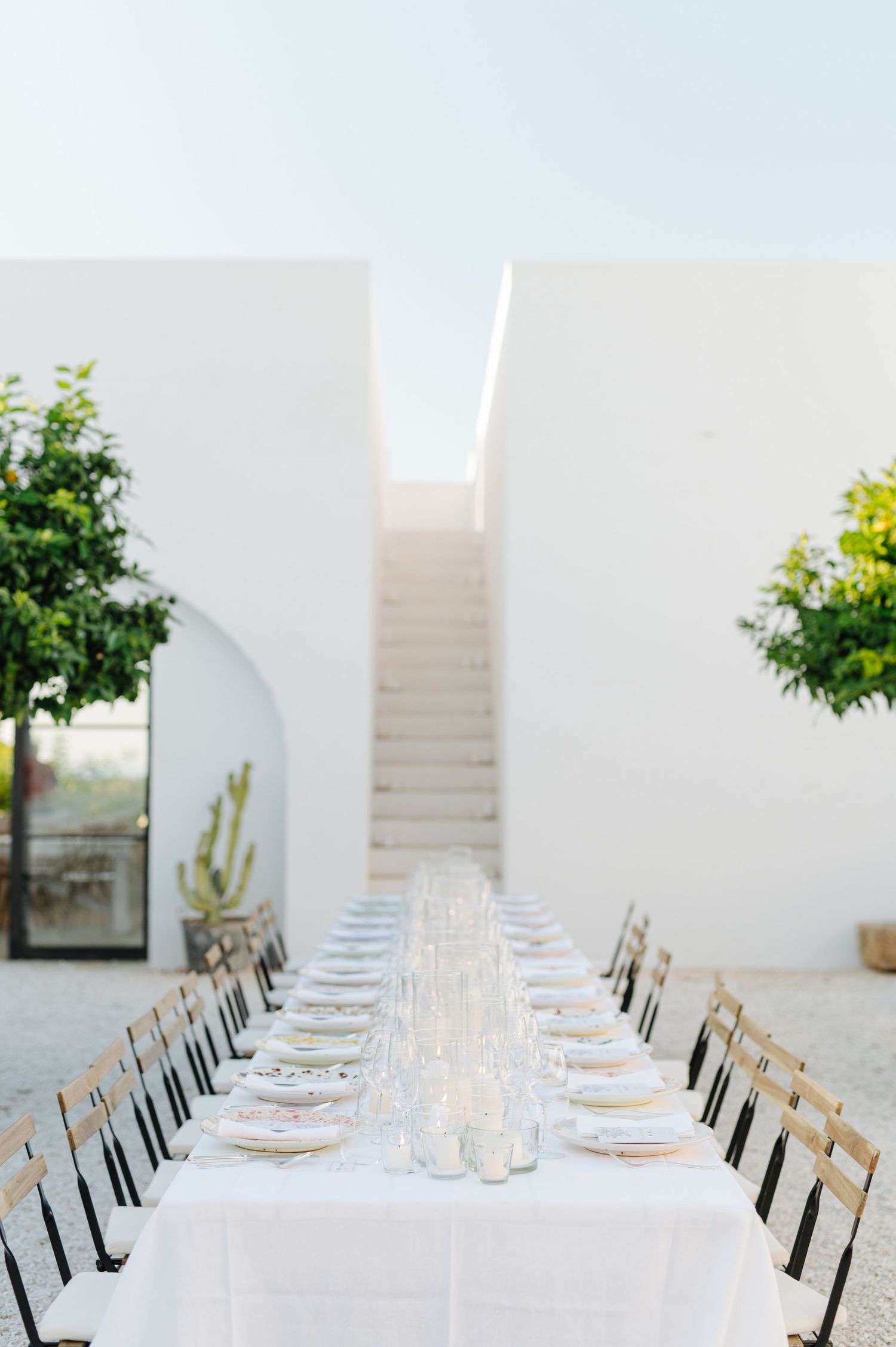 Elegant white dining table set with crystal glasses and black chairs in a minimalist Mediterranean setting.