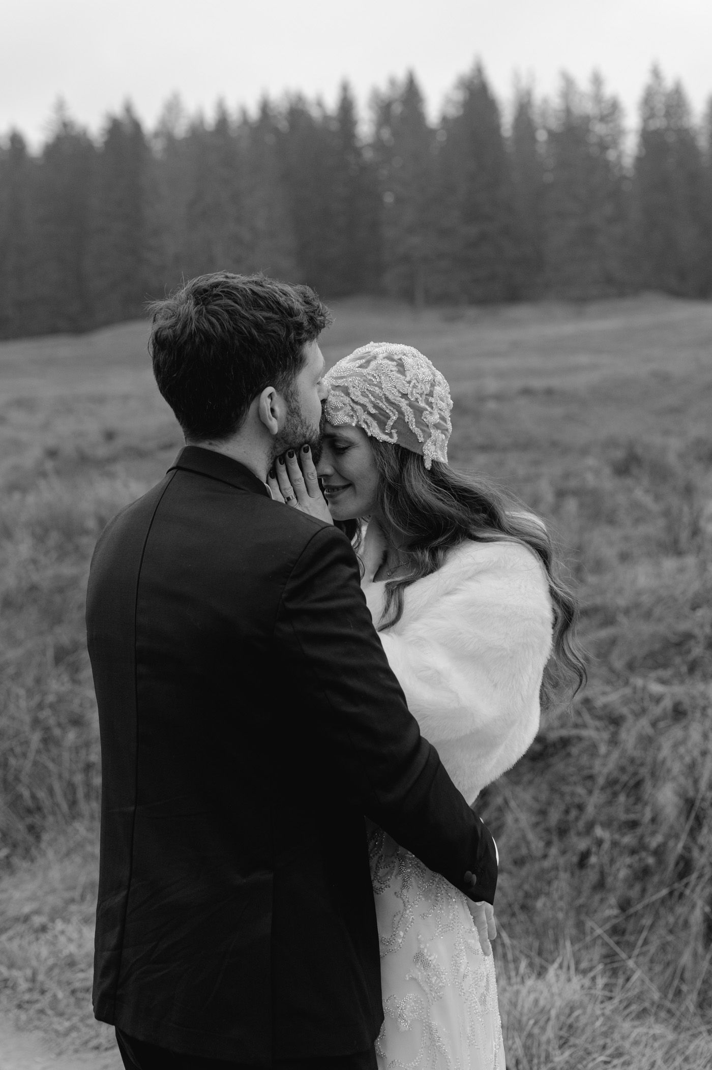 Black and white photo of a bride and groom embracing in a field, with pine trees in the background.