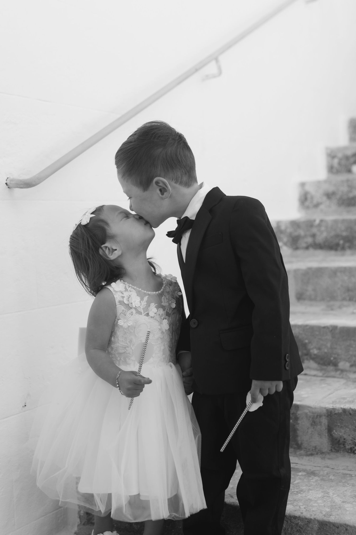 Two young children dressed formally kiss on stairs, boy in tuxedo and girl in white dress, black and white photo.