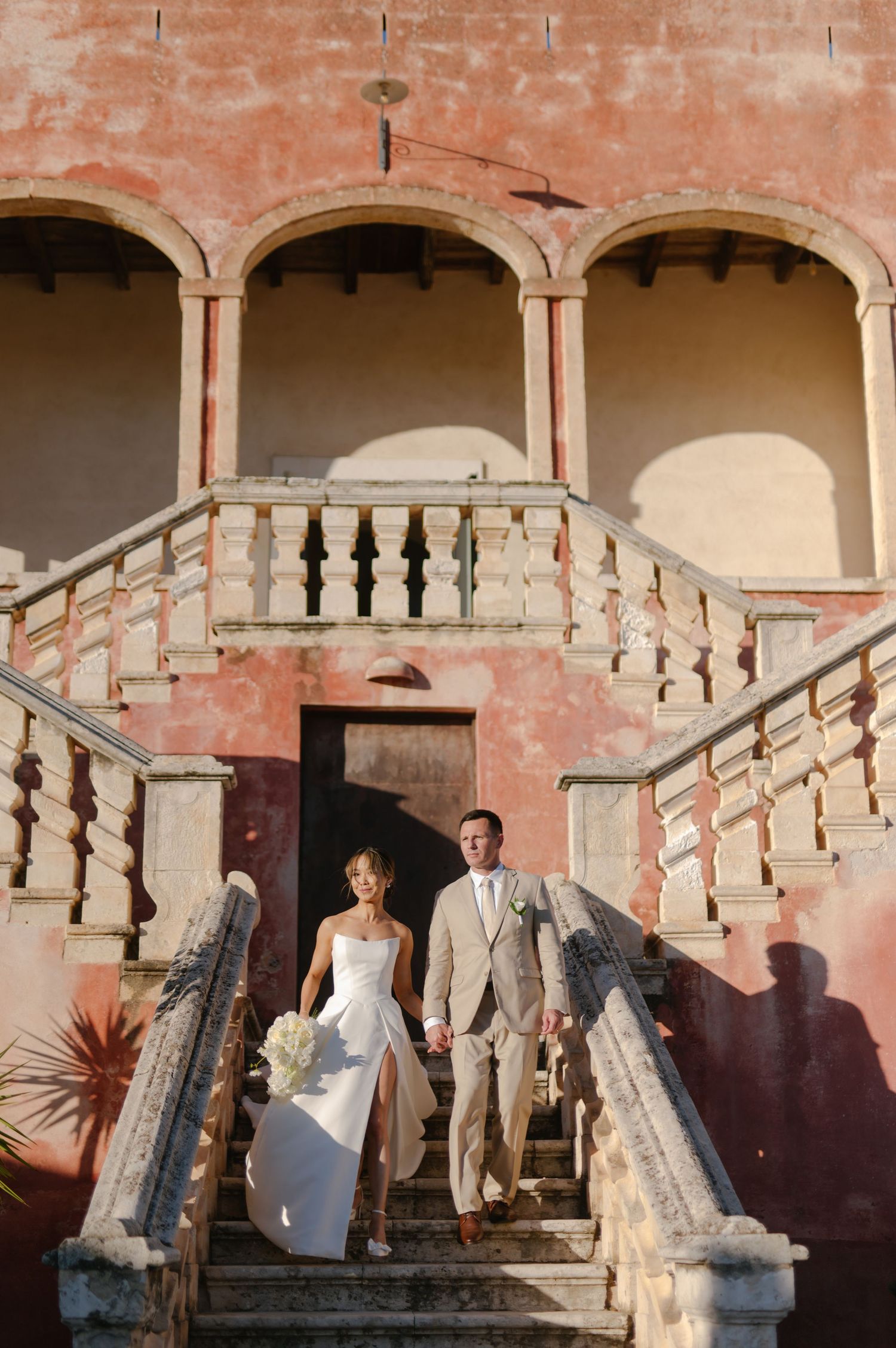 A couple descends an elegant stone staircase at a Mediterranean-style villa with pink walls and arched architecture.