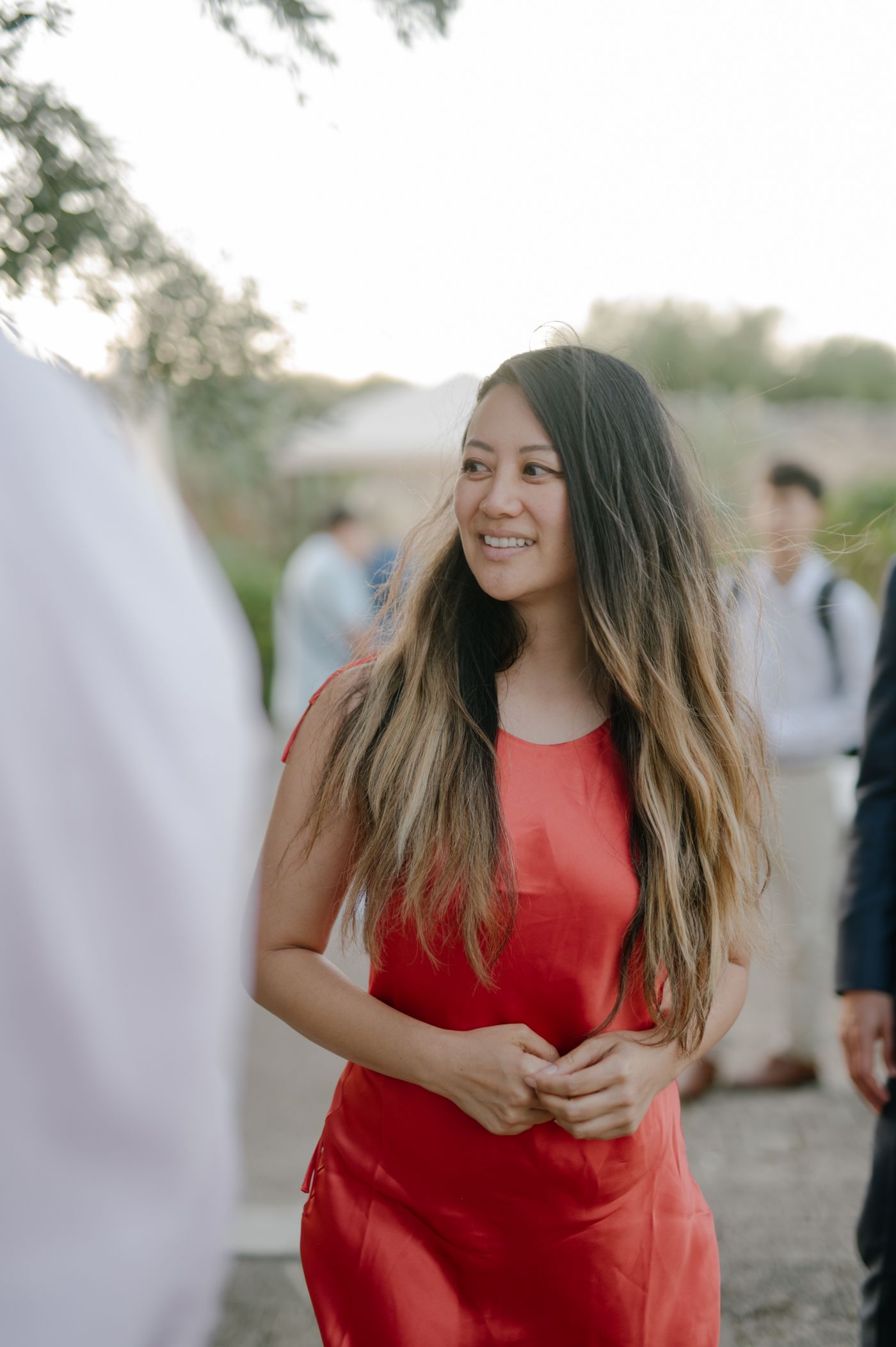 A person in a bright red sleeveless dress poses outdoors with long wavy hair against a natural background.