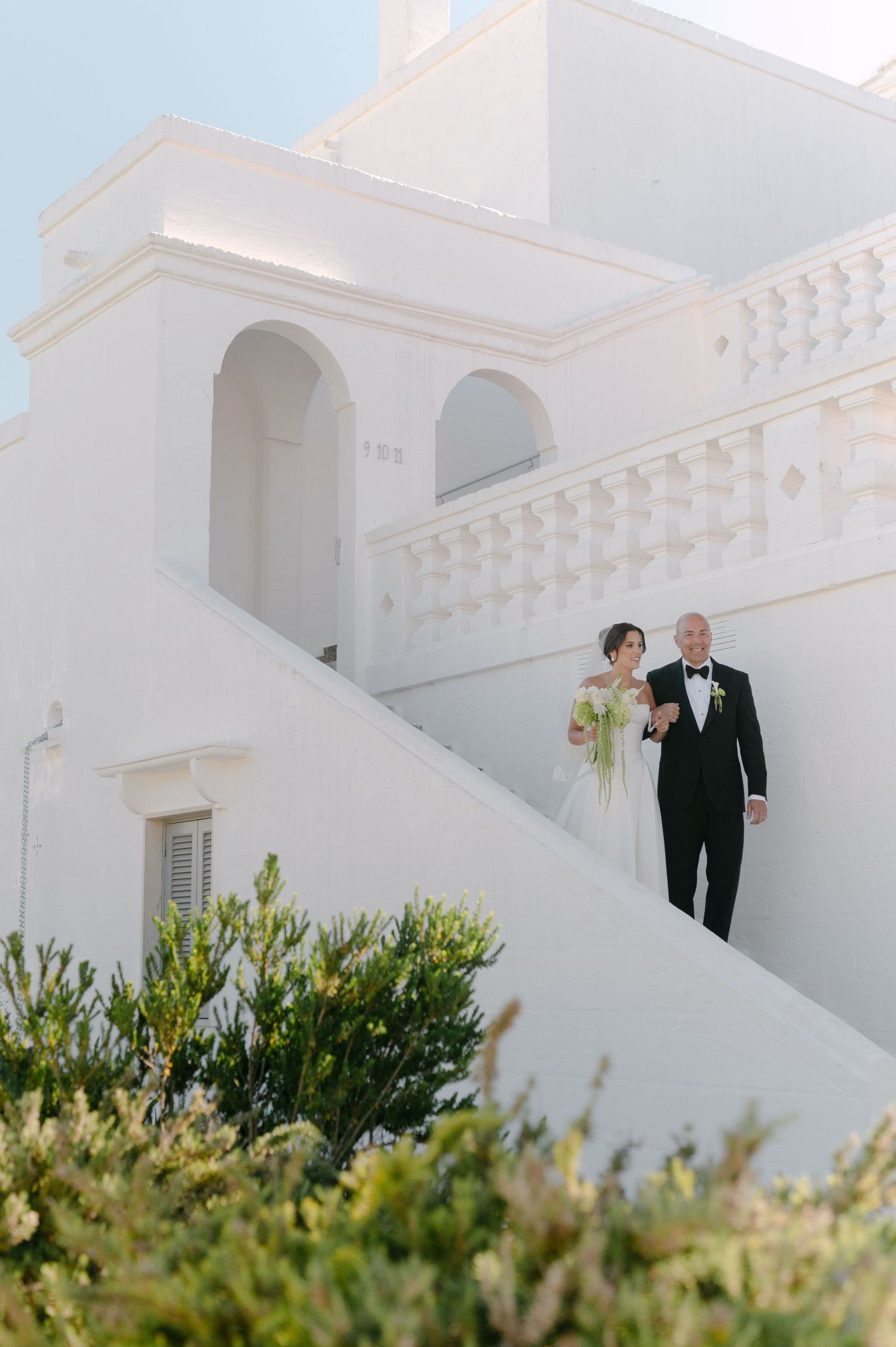 Bride and groom descend white staircase of elegant Mediterranean venue, bride holding green bouquet.