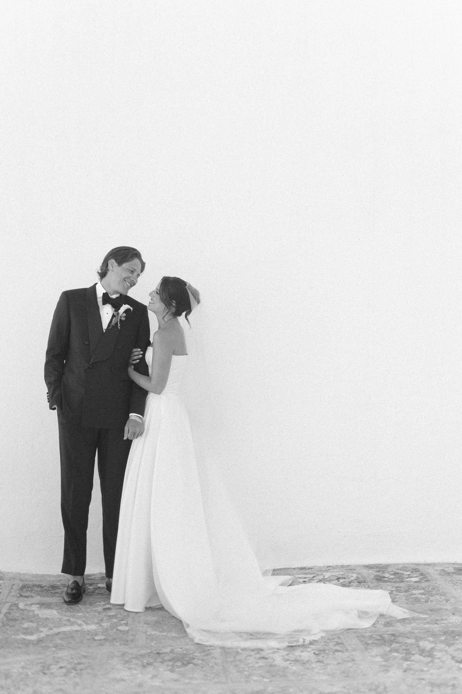 Black and white wedding photo of bride and groom standing together against a white wall in elegant attire.