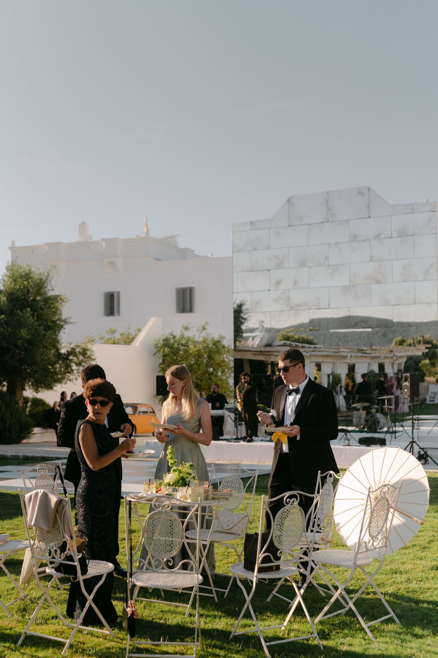 Elegant outdoor wedding reception on a green lawn with white iron chairs, guests in formal attire, and white Mediterranean buildings.
