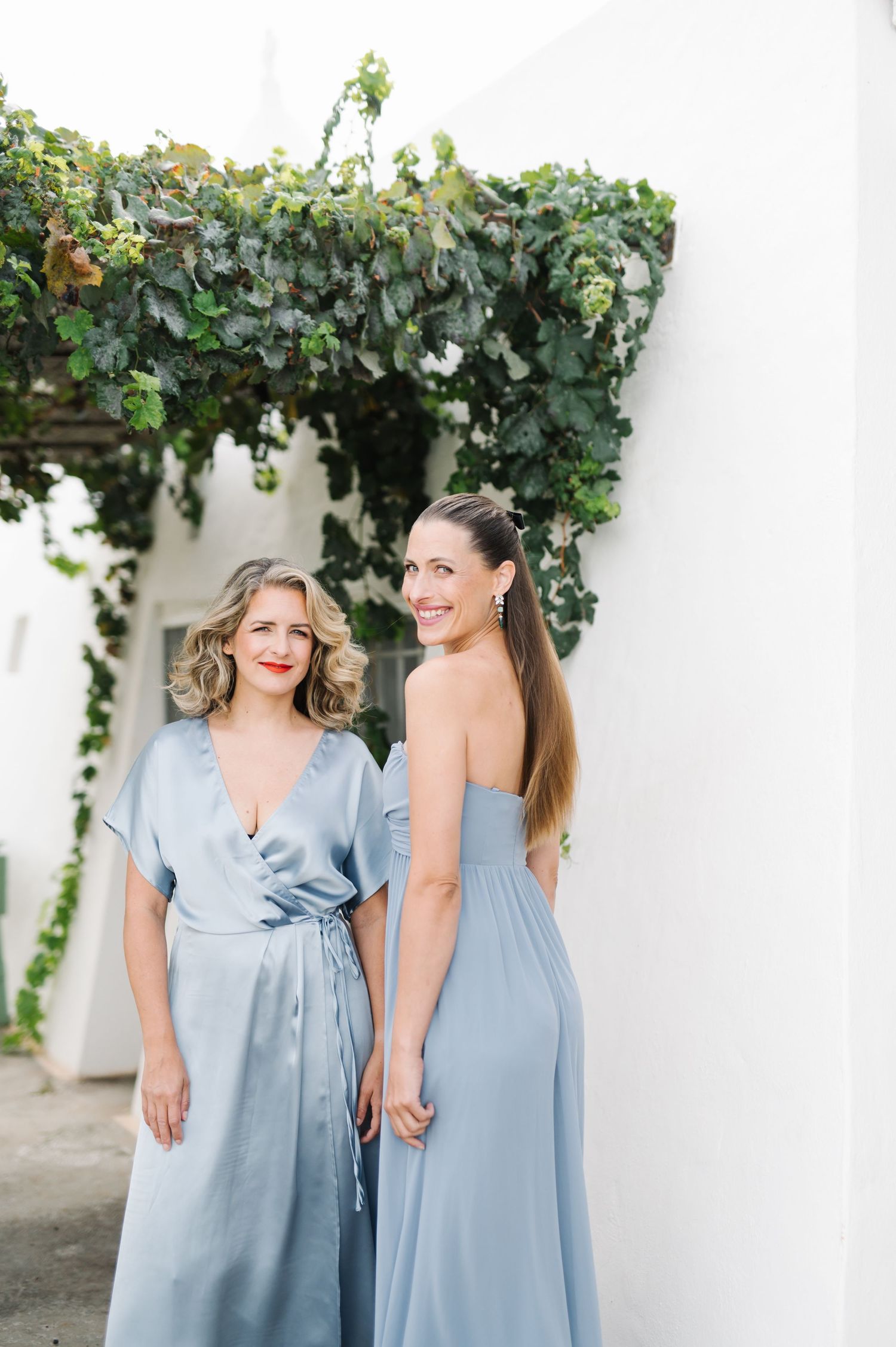 Bridesmaids in soft blue dresses posing against white wall with green vines.