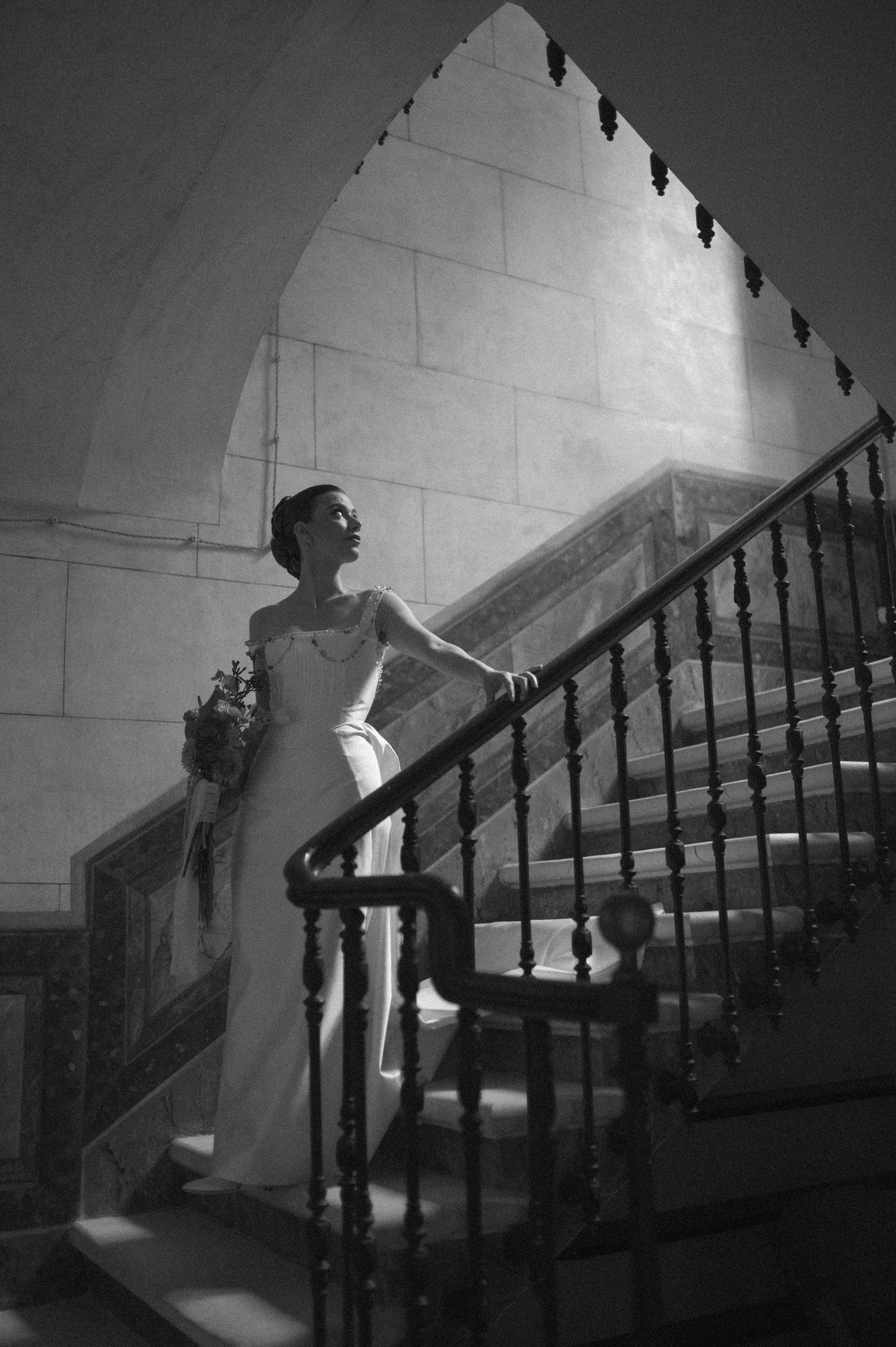 A black and white photograph of someone in a wedding dress ascending a grand staircase with an arched ceiling above.