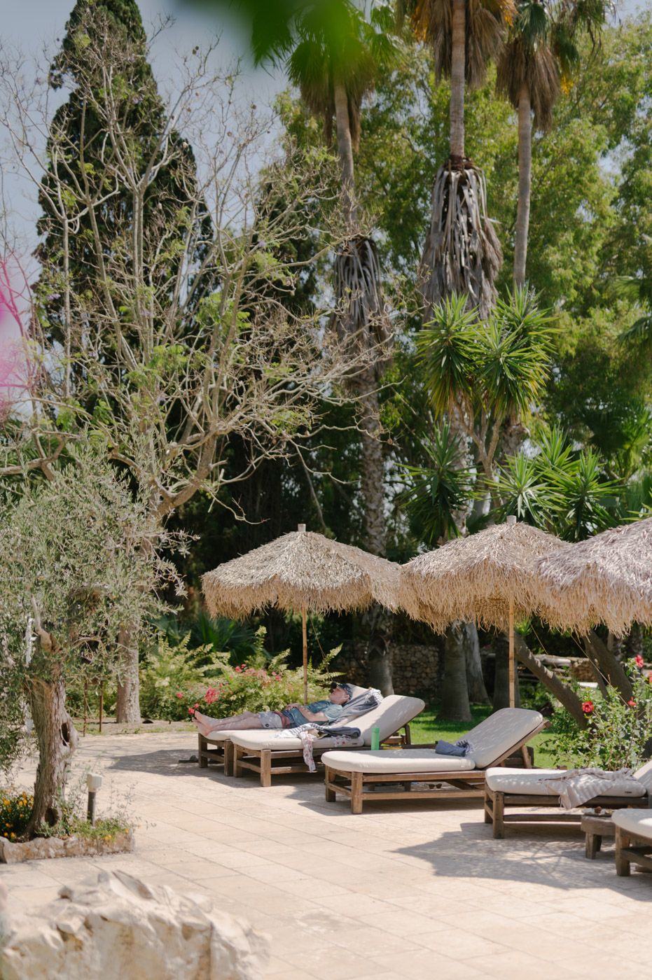Beachfront lounge area with thatched umbrellas and wooden sun beds surrounded by palm trees.