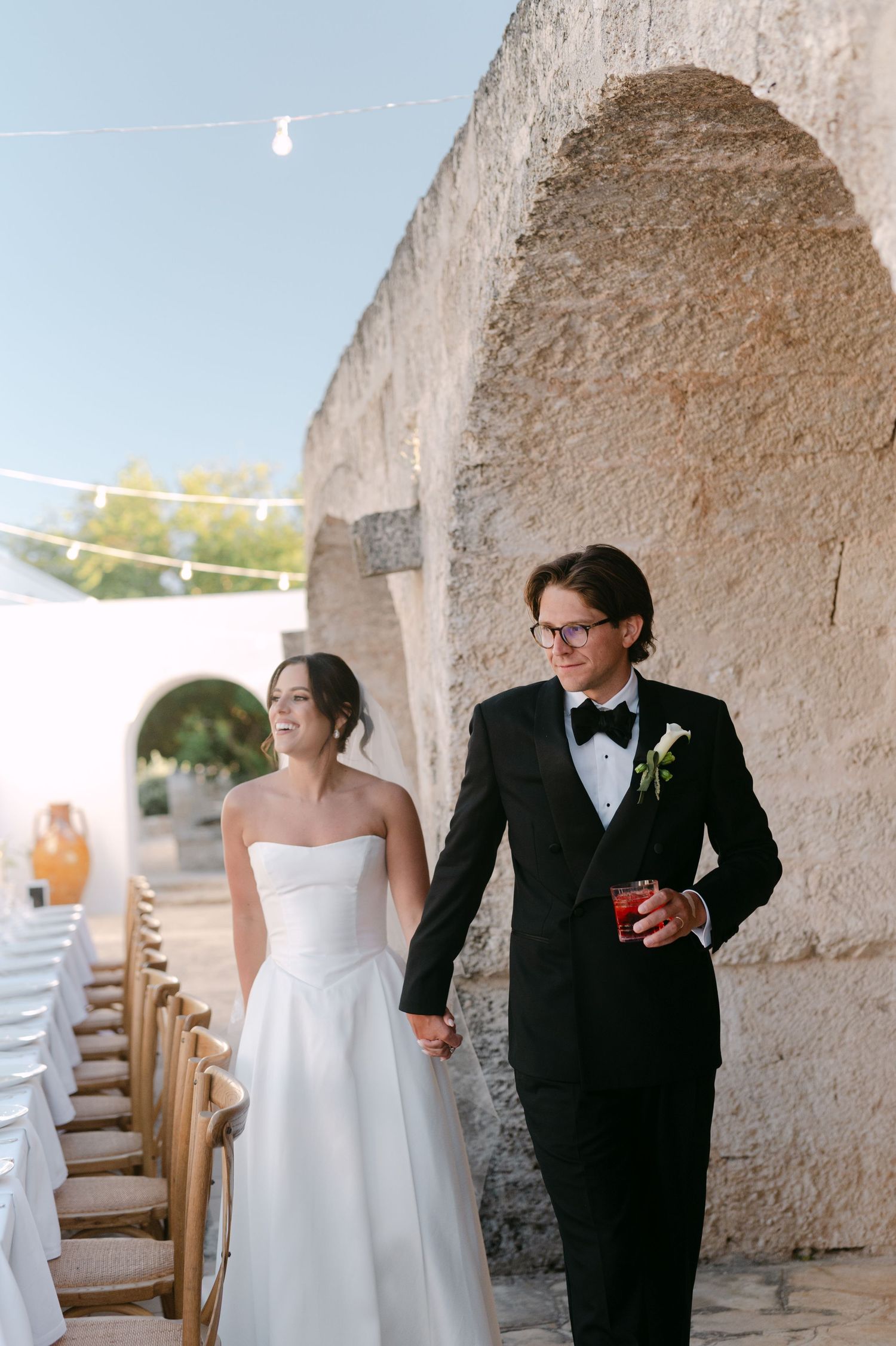A bride and groom walk hand-in-hand past a stone archway at an outdoor wedding venue.