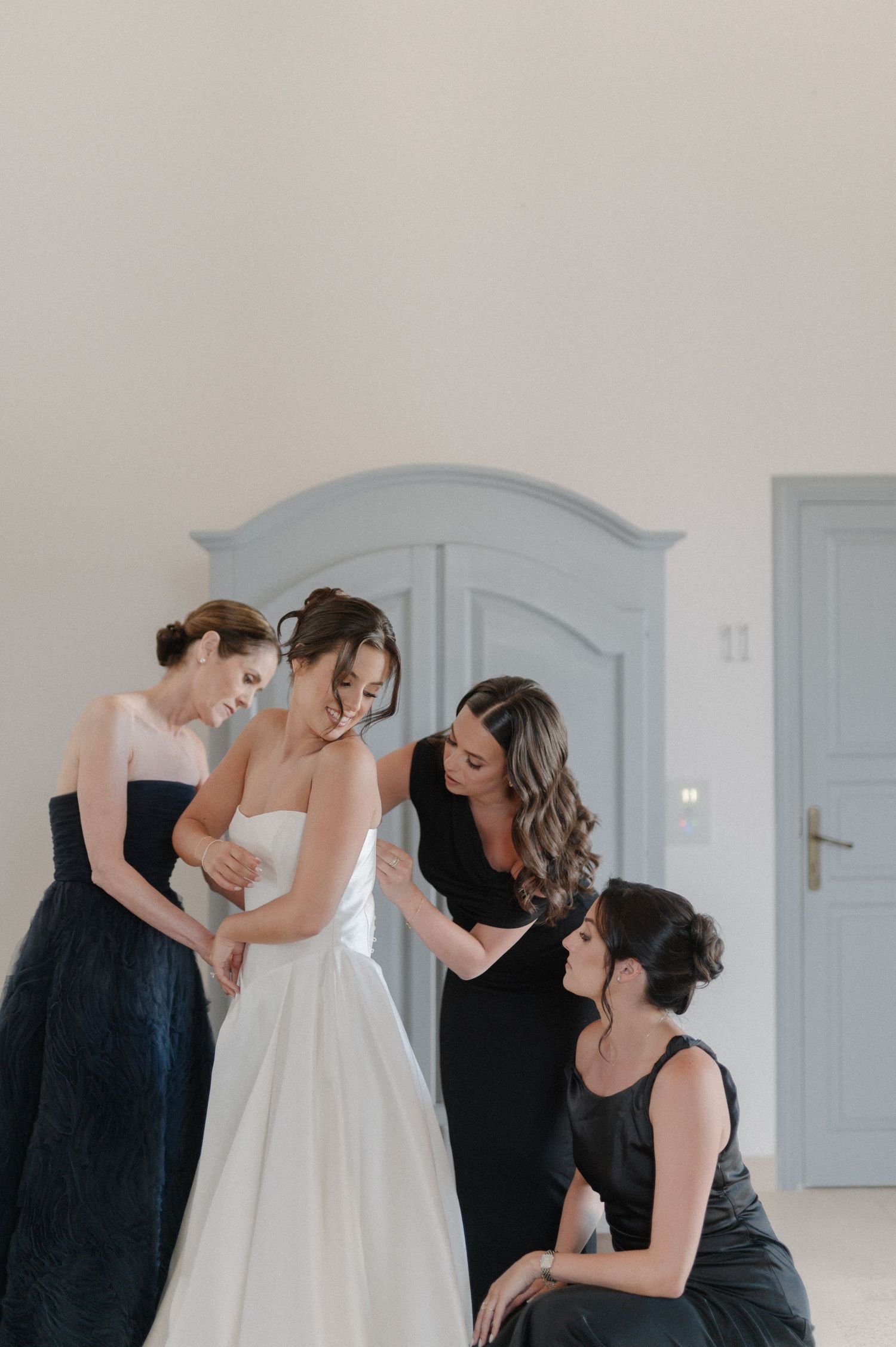Bride being helped into her white wedding gown by three bridesmaids in elegant dark dresses in a bright room.