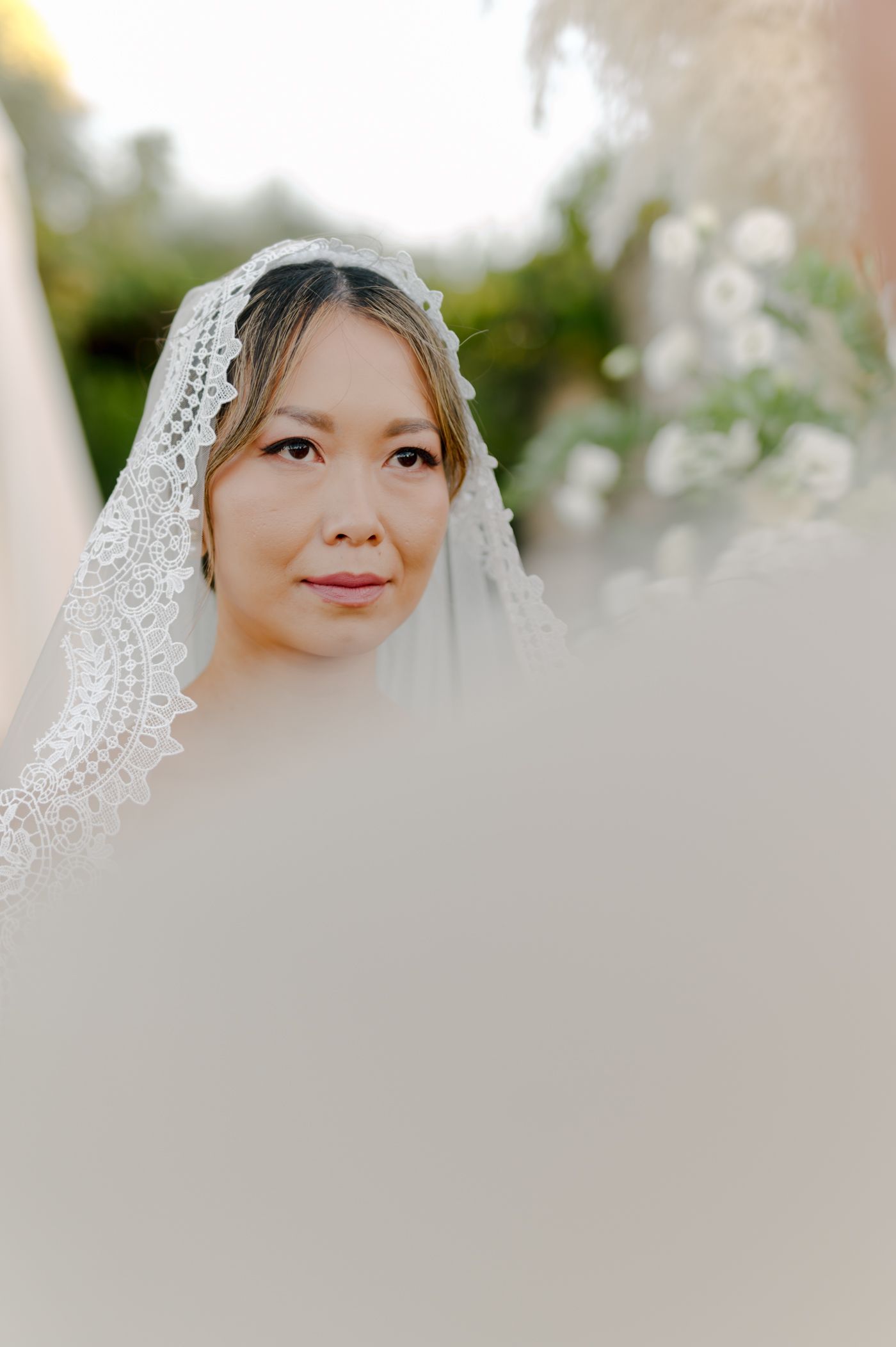 A bride wearing a delicate lace veil poses for a soft-focus portrait outdoors with natural lighting.