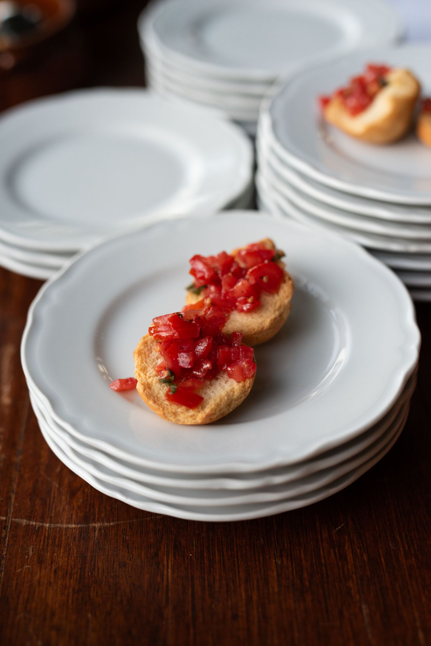 White plates stacked on dark wooden table with bruschetta topped with tomatoes and herbs.