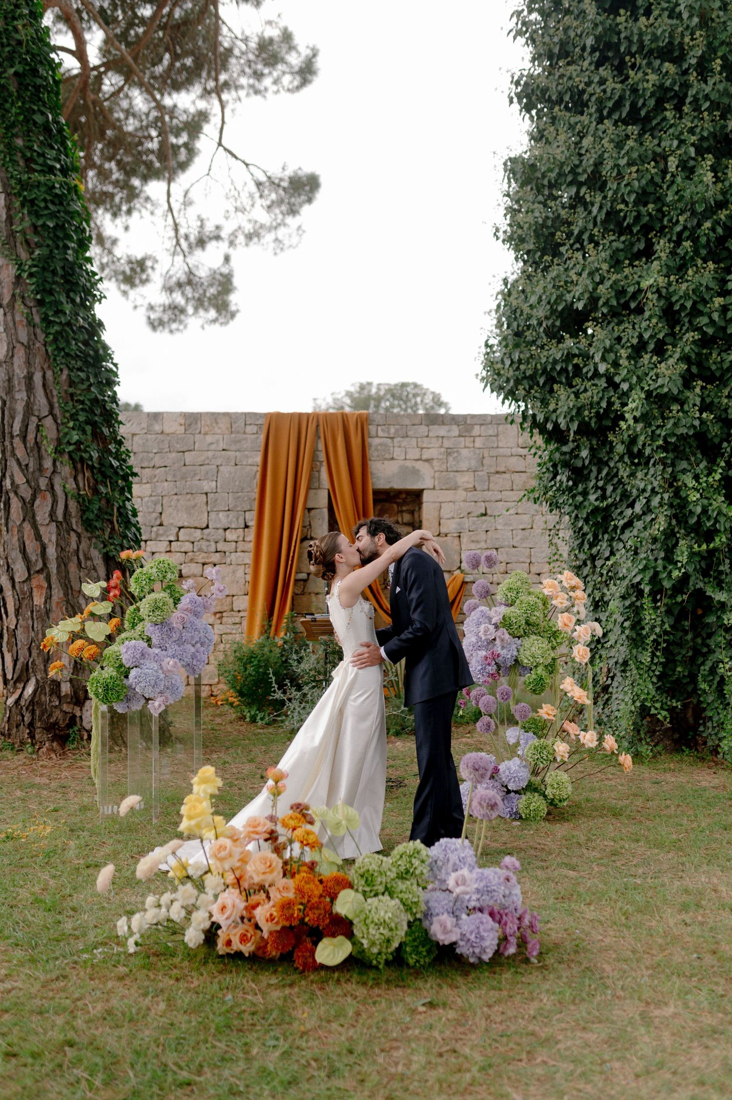 A couple shares an intimate moment in a garden setting with colorful floral arrangements against a stone wall.