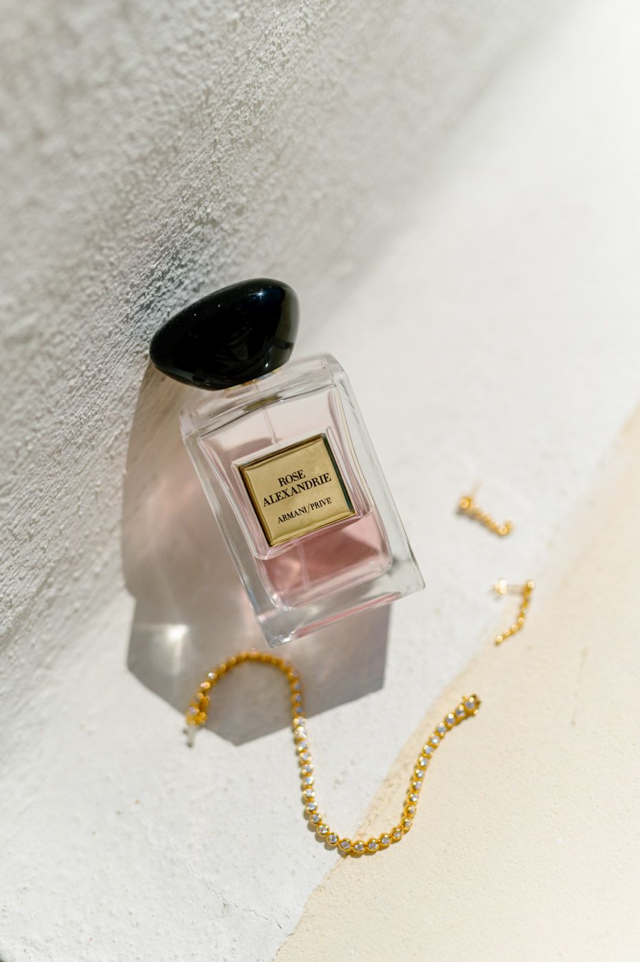 Perfume bottle and gold jewelry displayed on white textured surface with soft shadows.