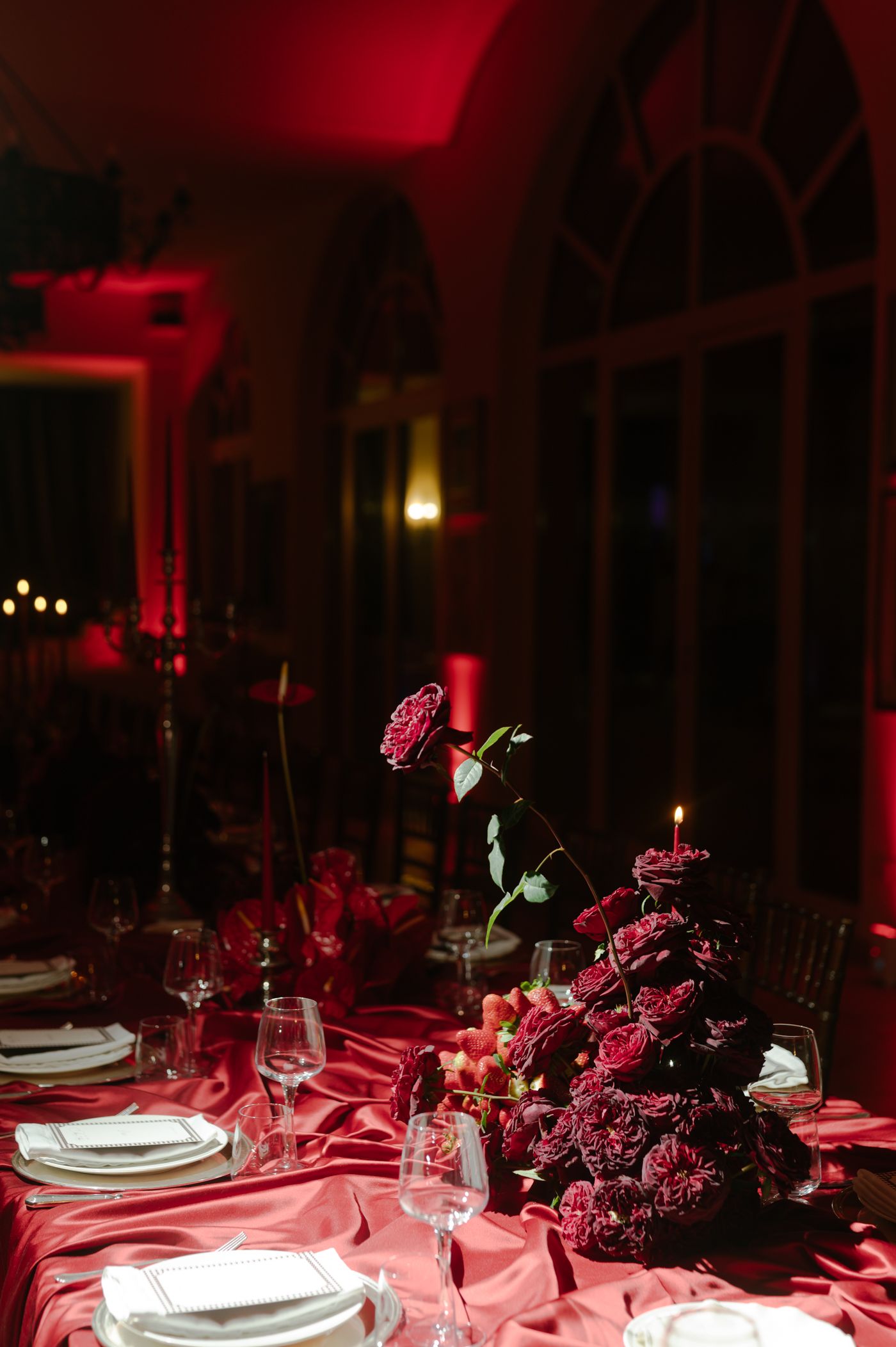Elegant dining table setup illuminated by dramatic red lighting with white place settings and dark floral centerpieces.