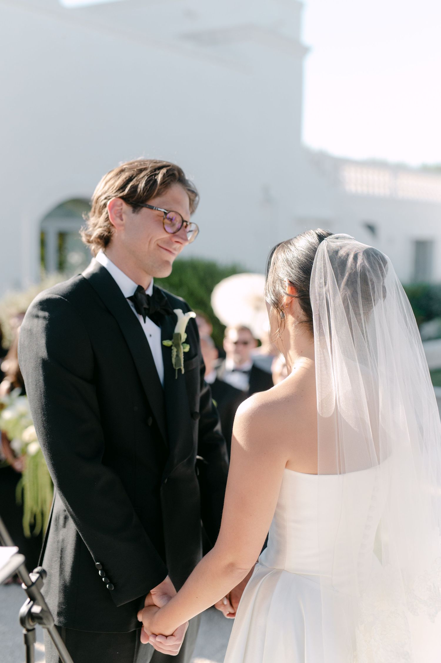 Groom in black tuxedo smiles at bride in white gown and veil while holding hands during outdoor wedding ceremony.