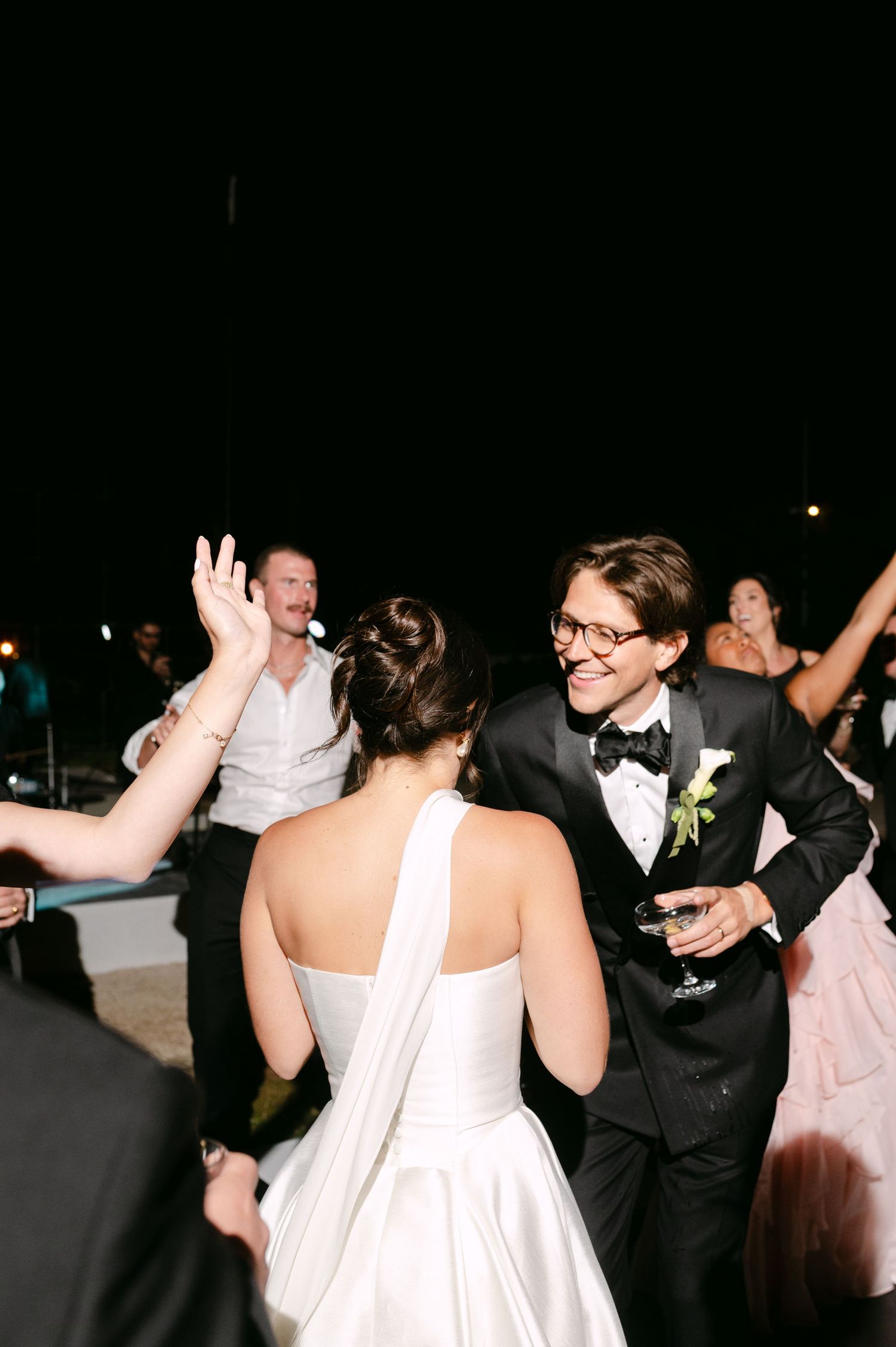 Bride and groom dancing joyfully at outdoor wedding reception at night, groom in black tuxedo with boutonniere.