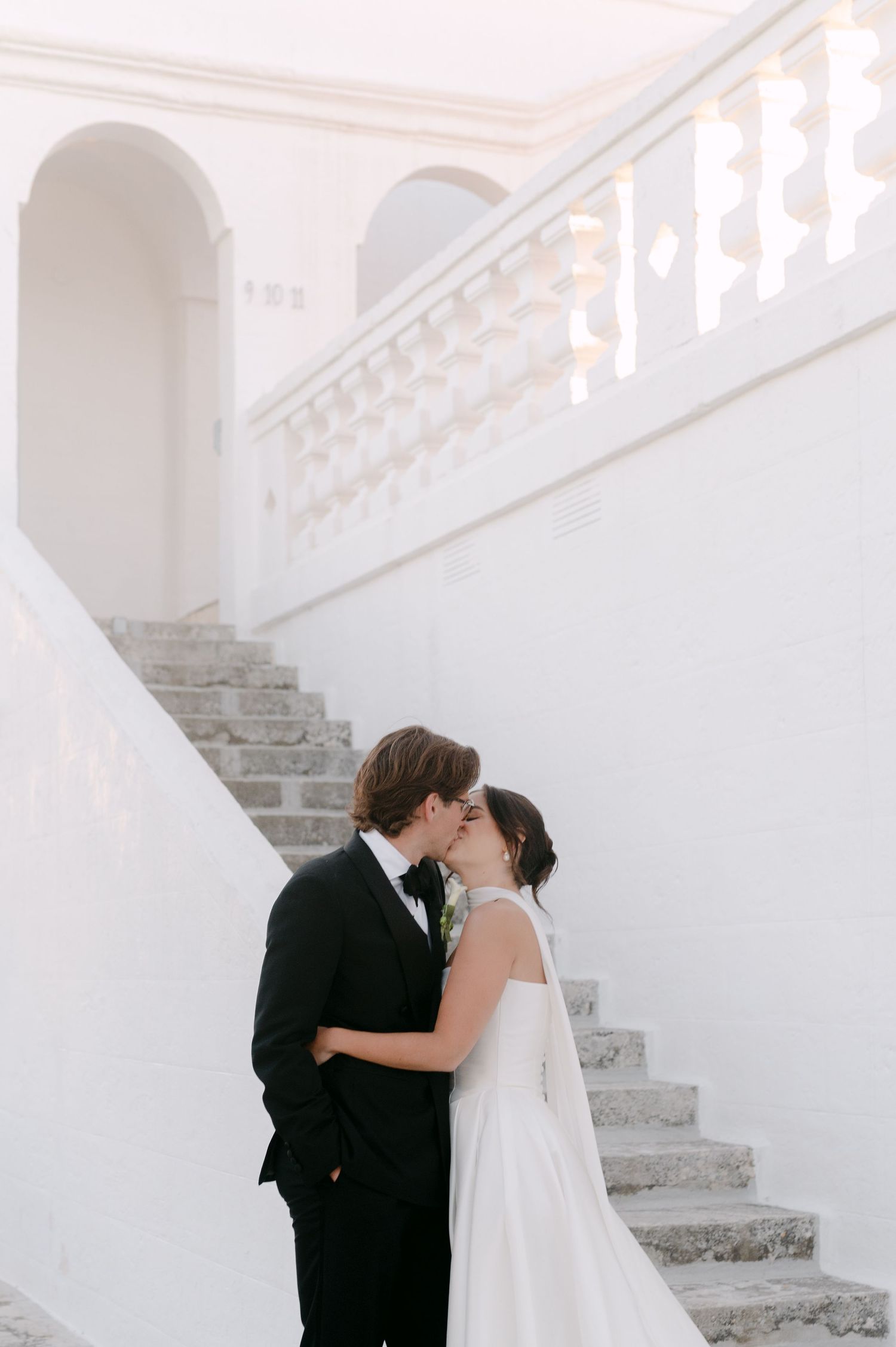Bride and groom share a romantic kiss beside elegant white marble stairs at a classic wedding venue.
