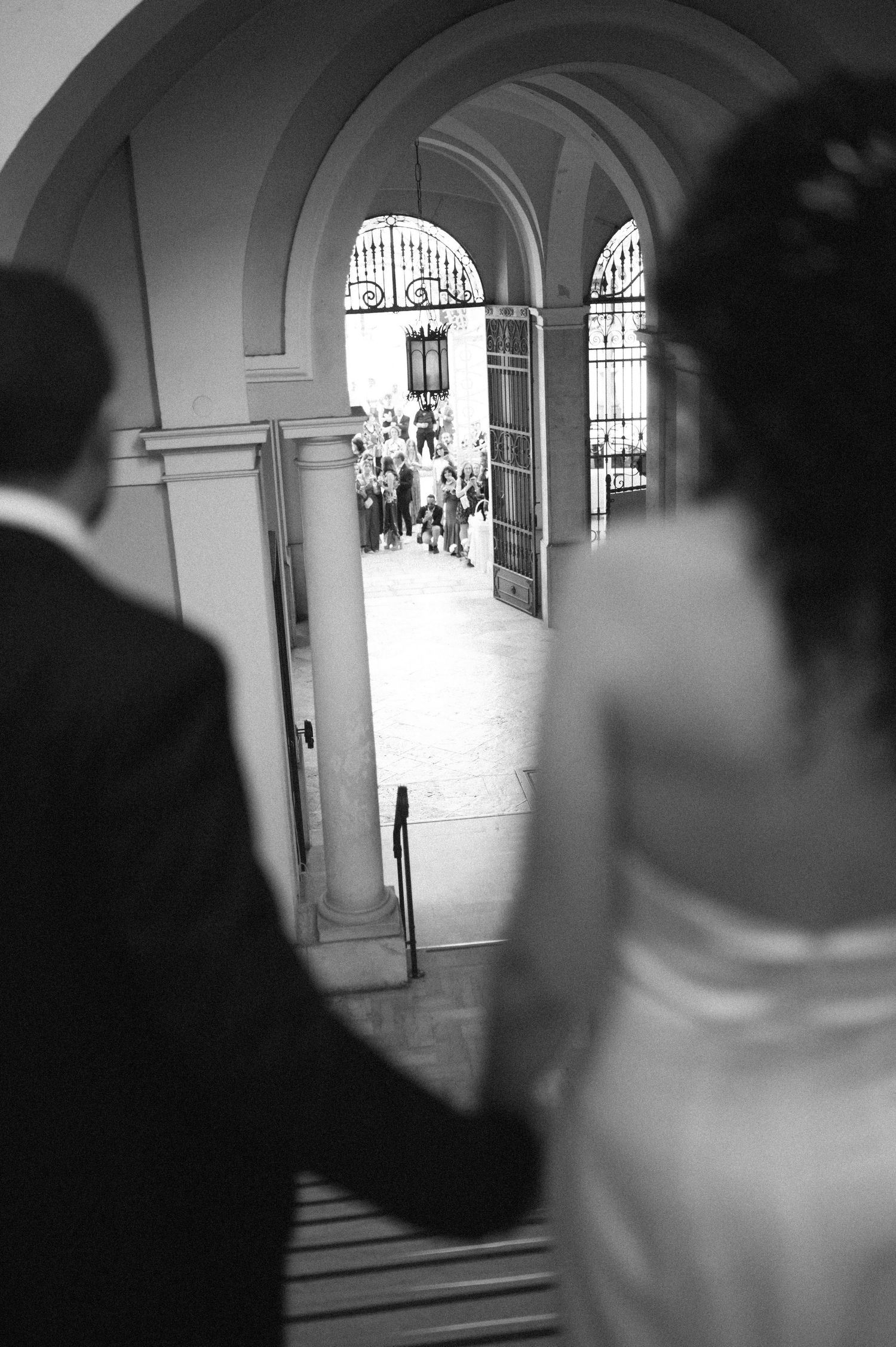 A black and white photograph showing a couple descending stairs through an arched doorway with guests visible beyond.