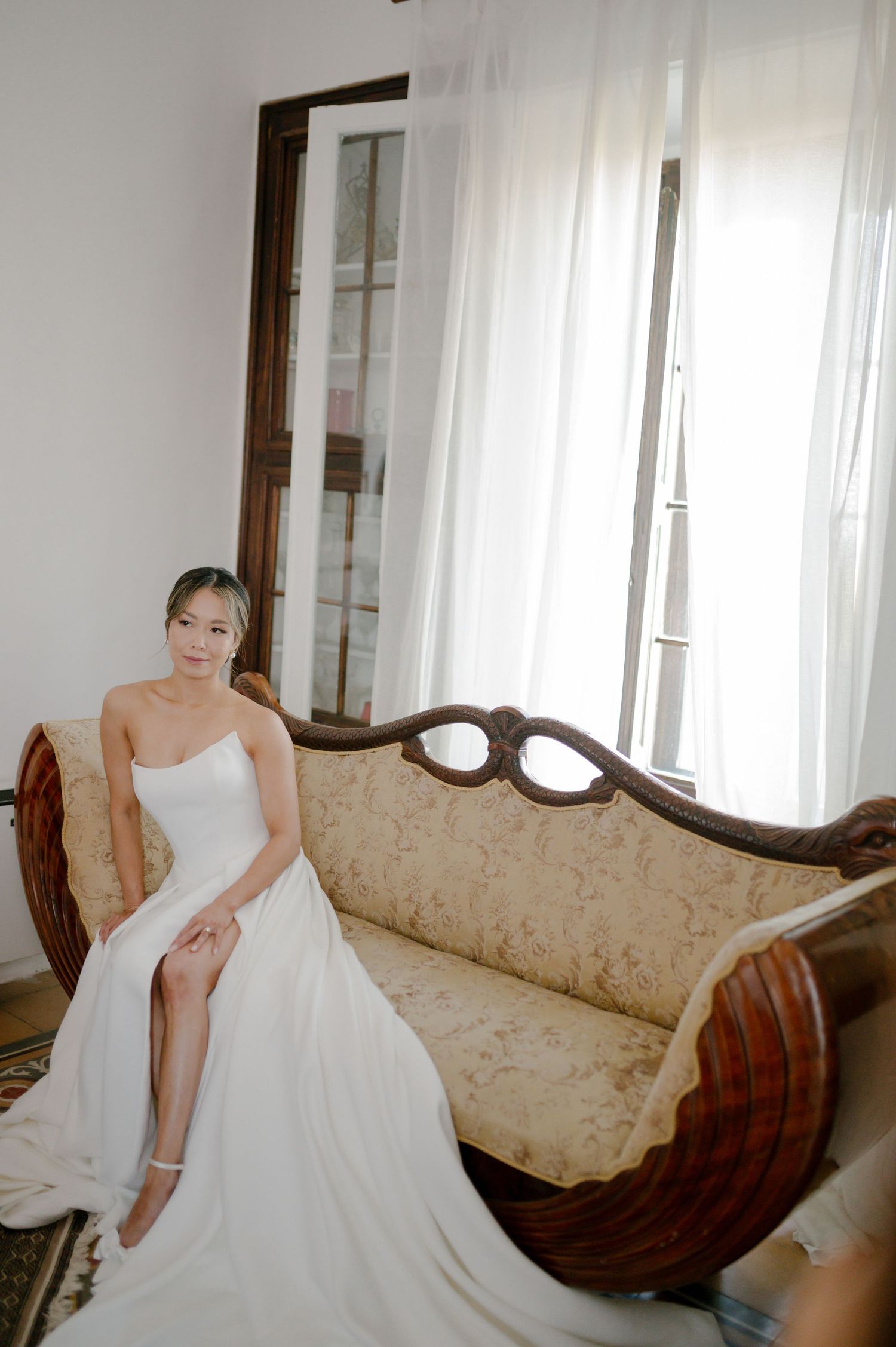 A figure in a flowing white wedding gown sits elegantly on a vintage beige sofa near sunlit windows.