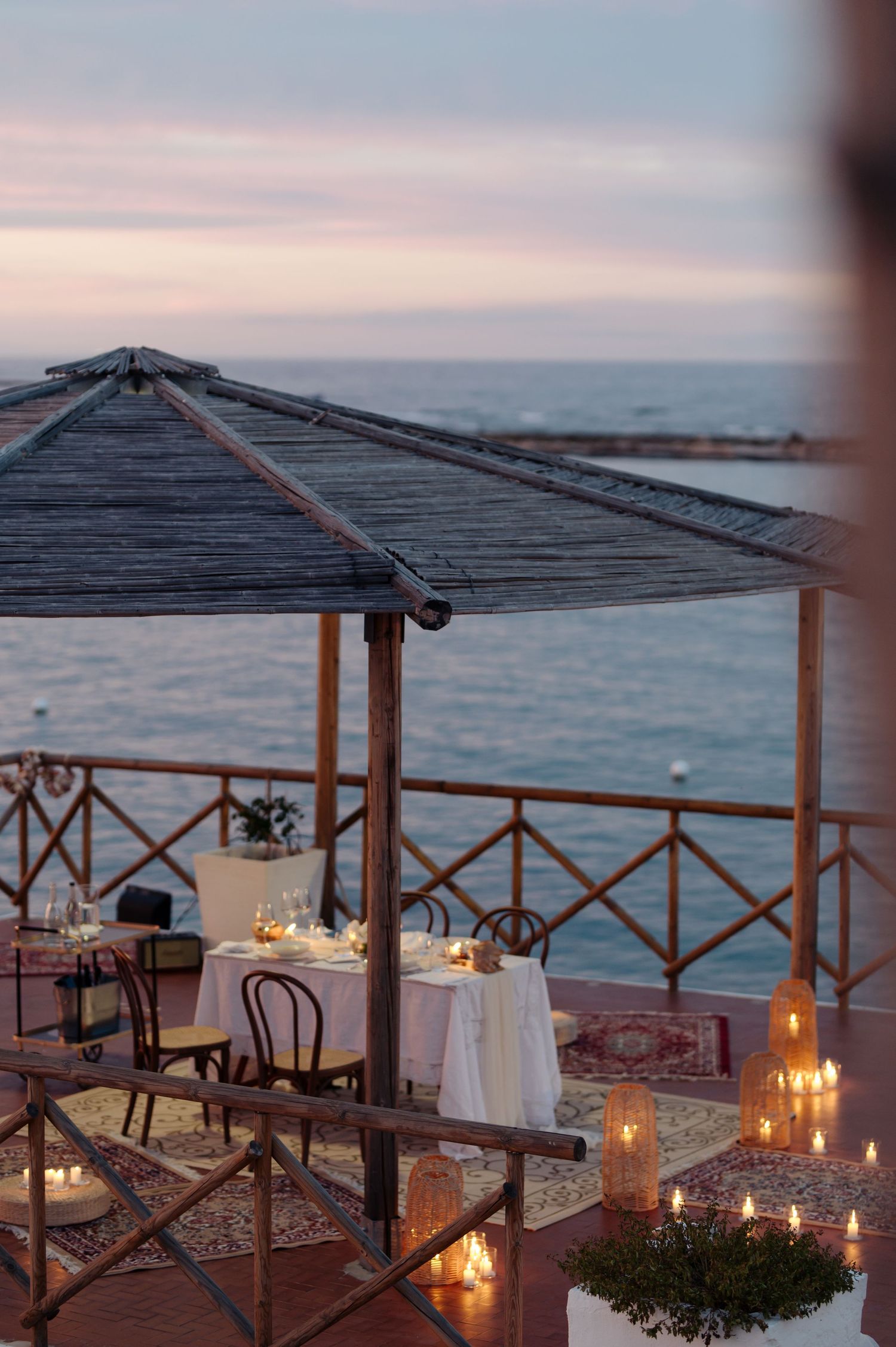 Romantic seaside dining setup with lantern-lit tables under wooden pergolas at sunset overlooking the ocean.