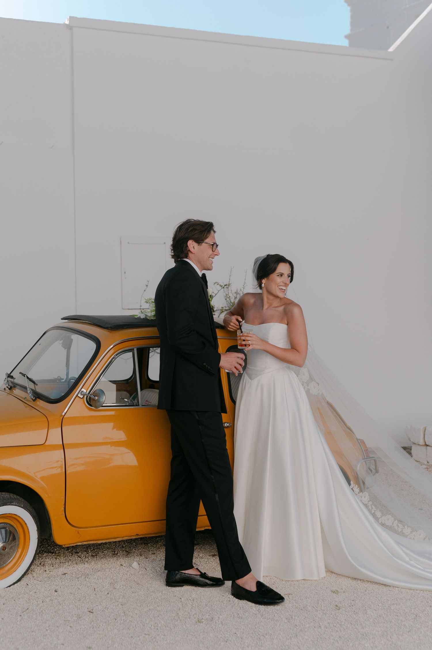 Bride and groom leaning against a vintage yellow Fiat 500 car on their wedding day in a sunny outdoor setting.
