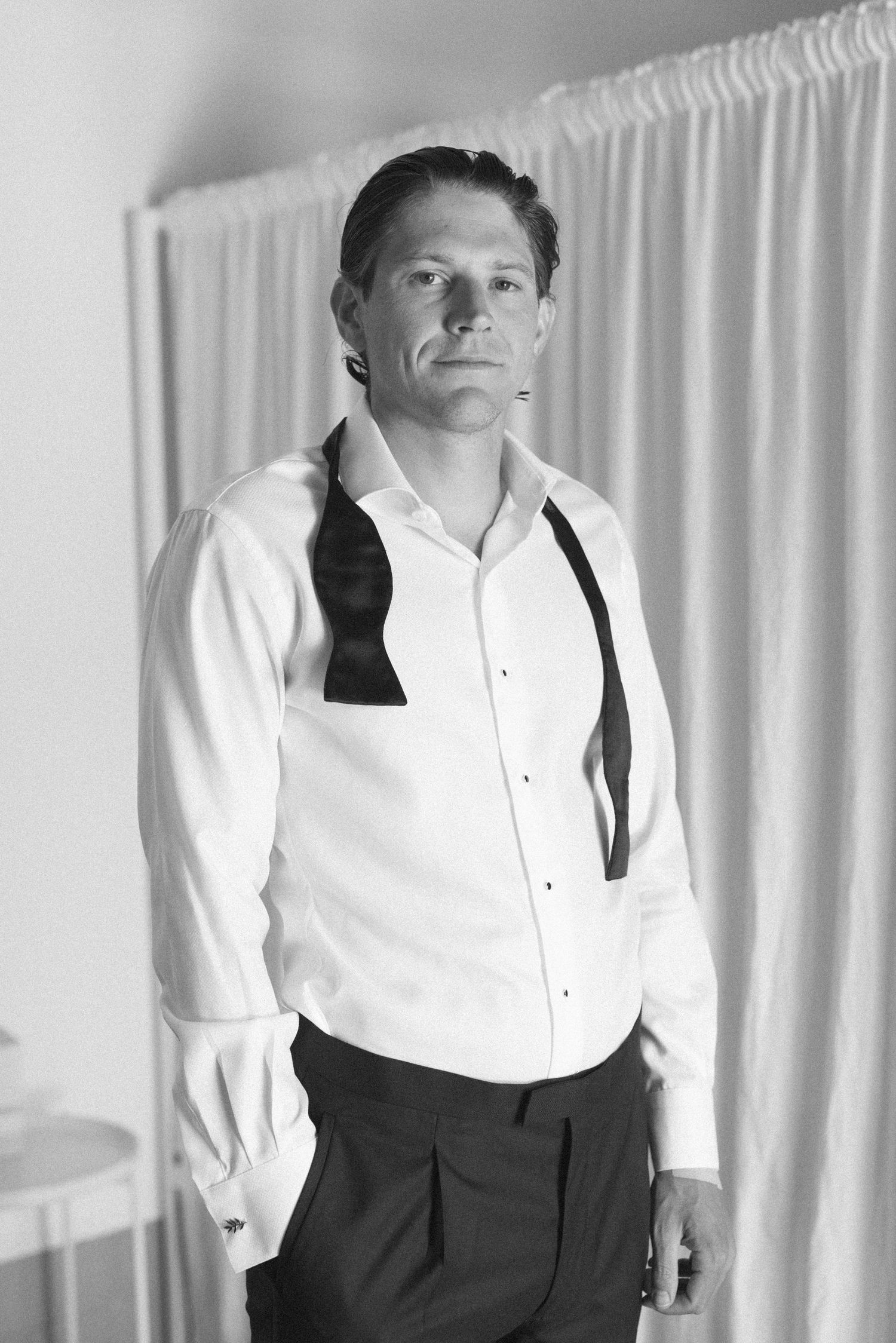 Black and white portrait of a man in a white dress shirt with bow tie and suspenders, posing indoors.