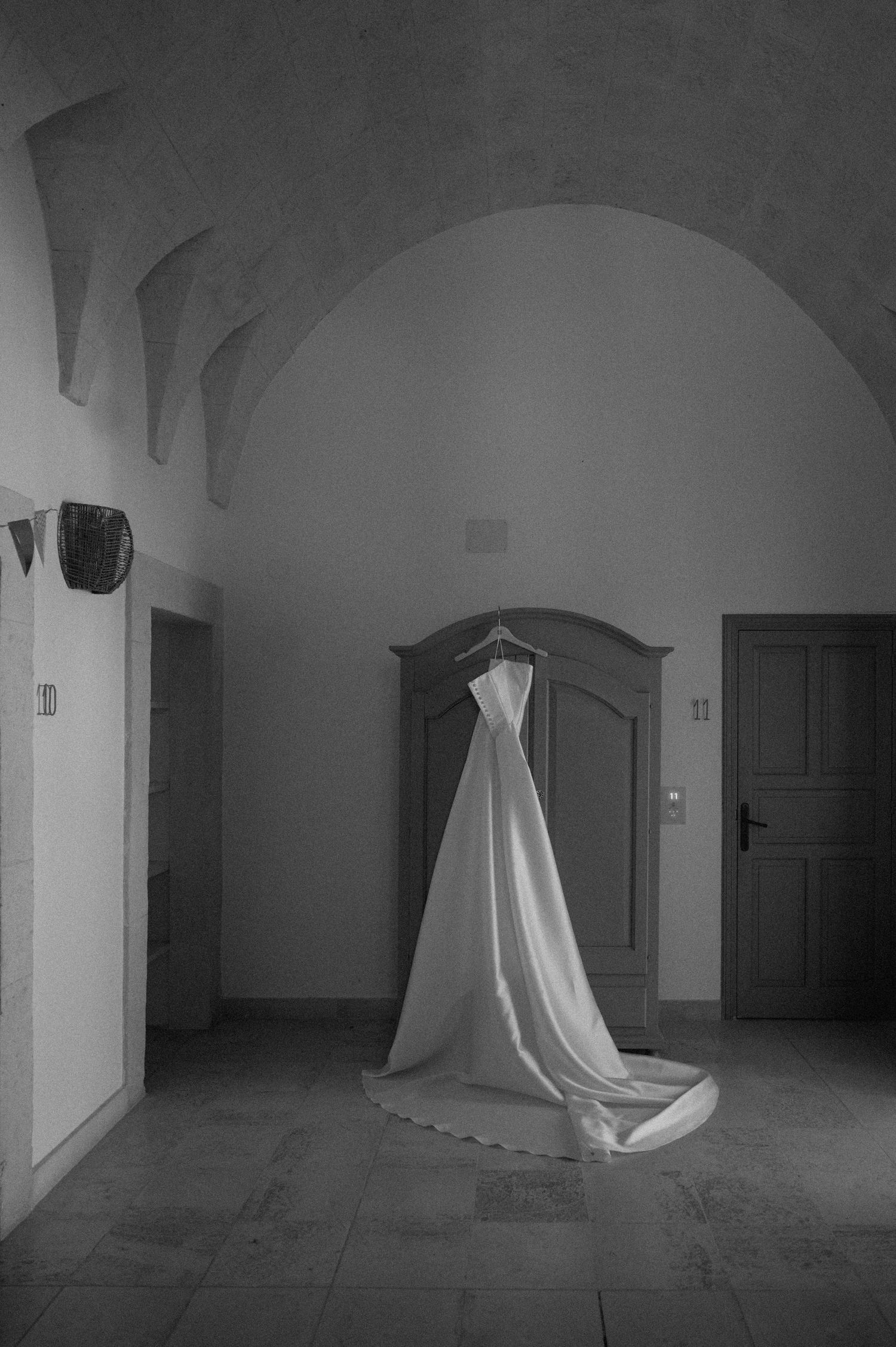 Black and white photo of an elegant wedding dress hanging in an arched stone hallway.