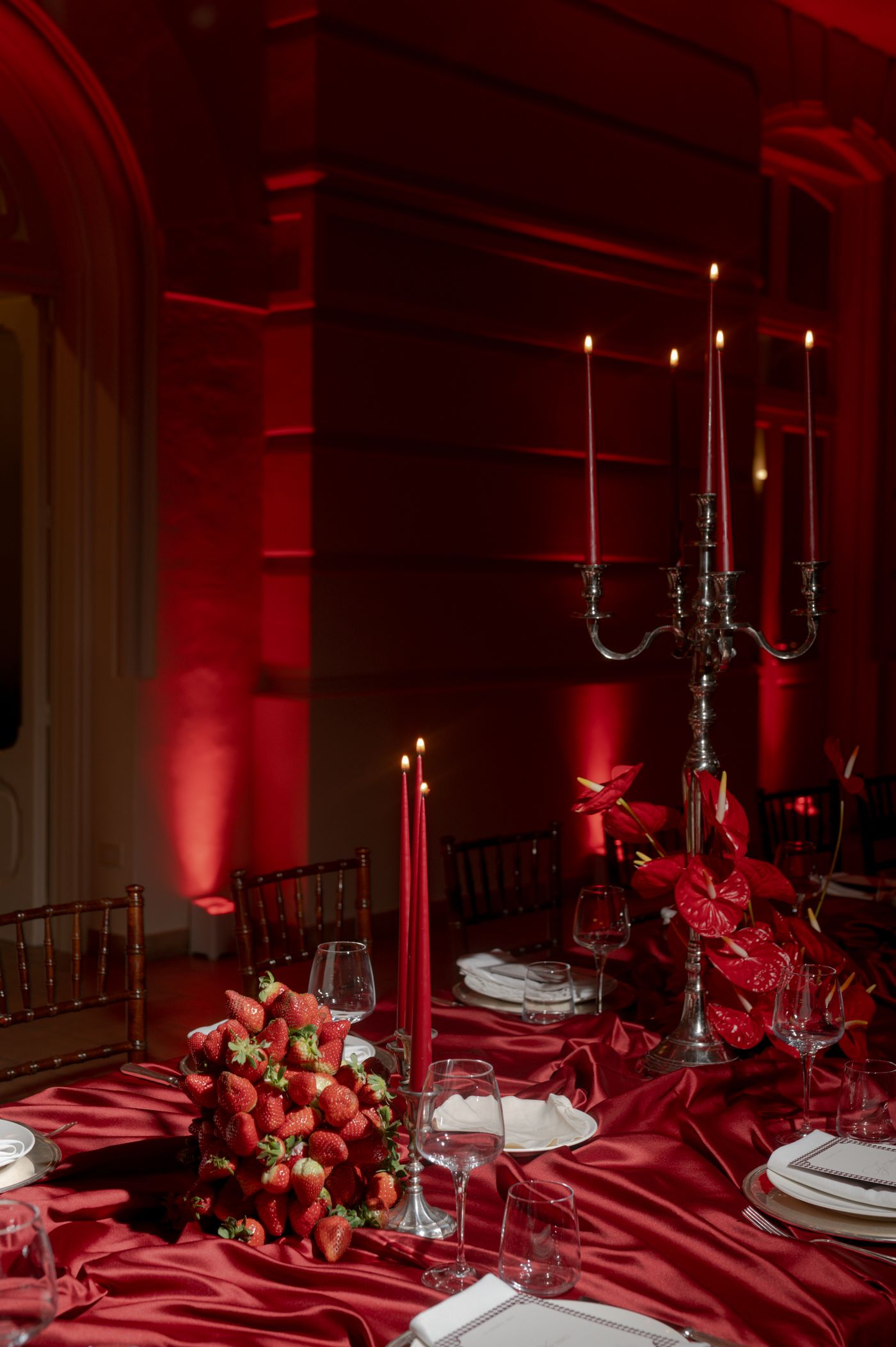Elegant red tablescape with silver candelabras, white plates, and fresh strawberry decorations on crimson satin tablecloth.